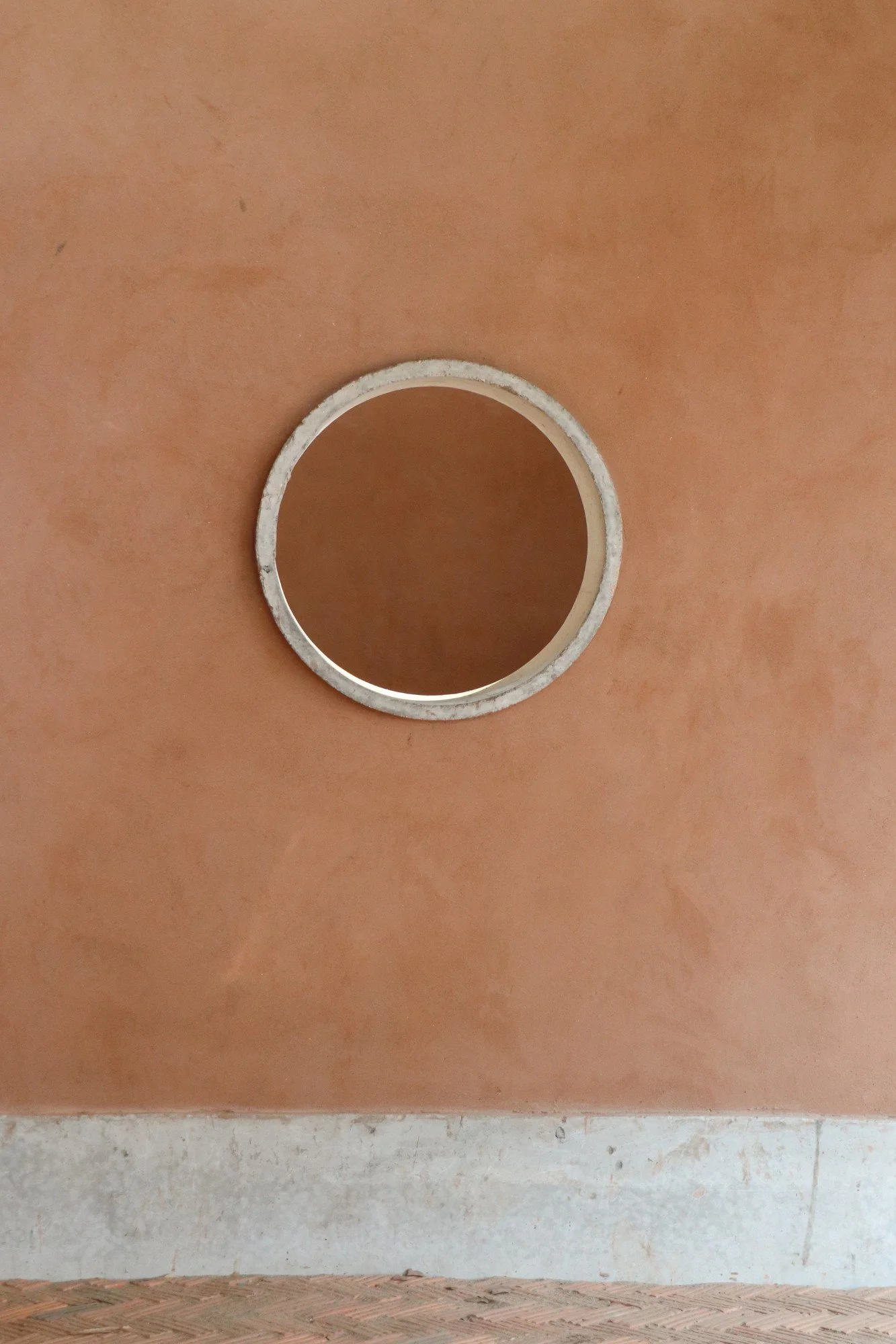 Ein runder Spiegel an einer orangefarbenen Wand, darunter eine weiße Wand und eine Bodenfliese.