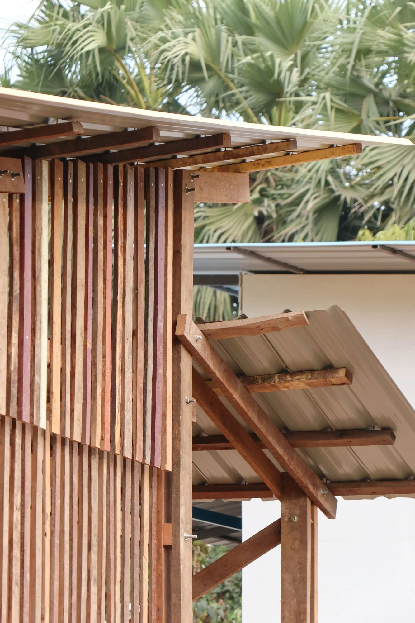 Ein Holzrahmen und eine Verkleidung aus Holzlatten, die vermutlich ein Balkon oder eine Terrasse bauen. Im Hintergrund sind Palmen zu sehen.