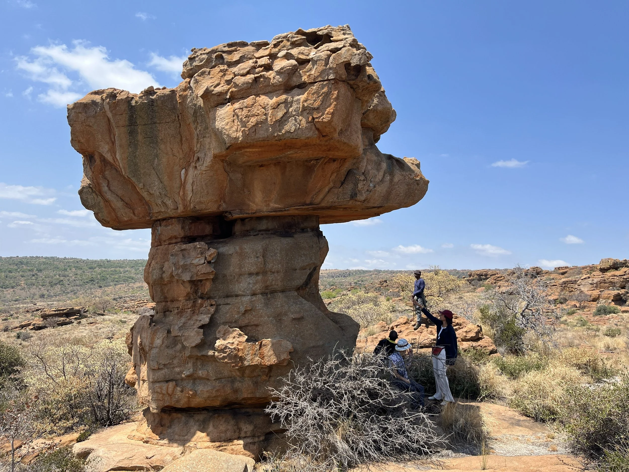 A large, balanced rock formation in a desert landscape with four people nearby, two standing and two sitting, under a partly cloudy sky.