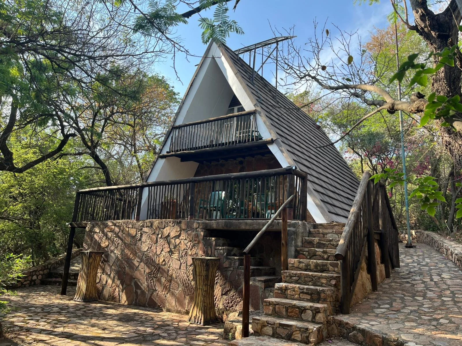 A small, A-frame house with a stone porch and black railings, surrounded by trees, under a partly cloudy sky.