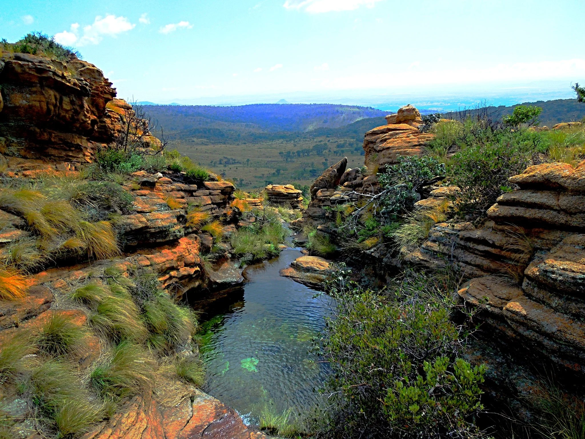 Rugged rocky landscape with patches of grass and shrubs, a small water pool in foreground, distant hills under cloudy sky.