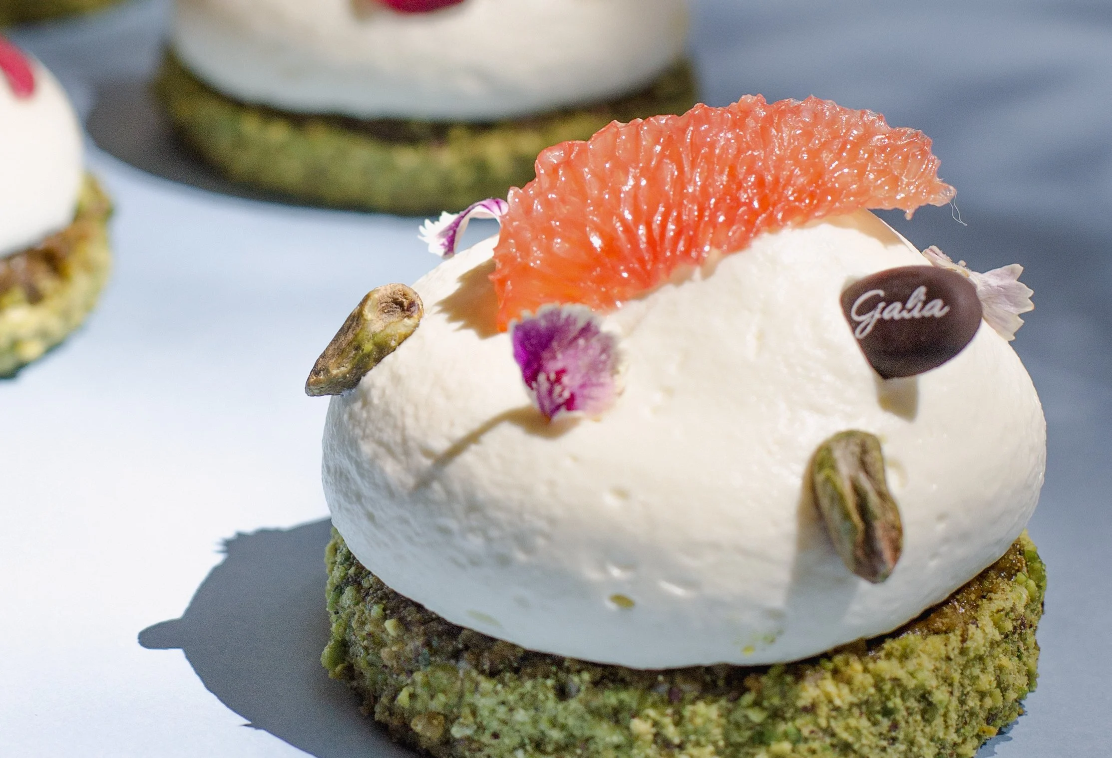 Postre con base de pistaches triturados, crema blanca decorada con una rebanada de naranja, pétalos de flores y pedacitos de pistache.