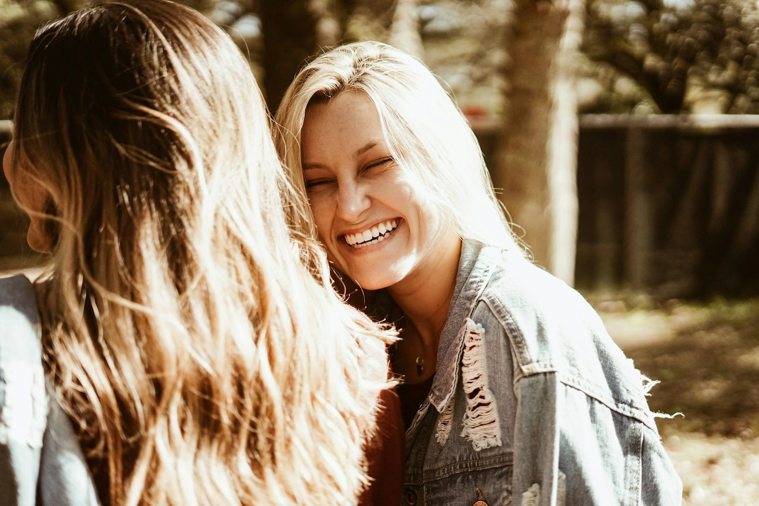 Two women sharing a joyful moment outdoors, one with blonde hair and the other with brown hair, smiling and laughing.