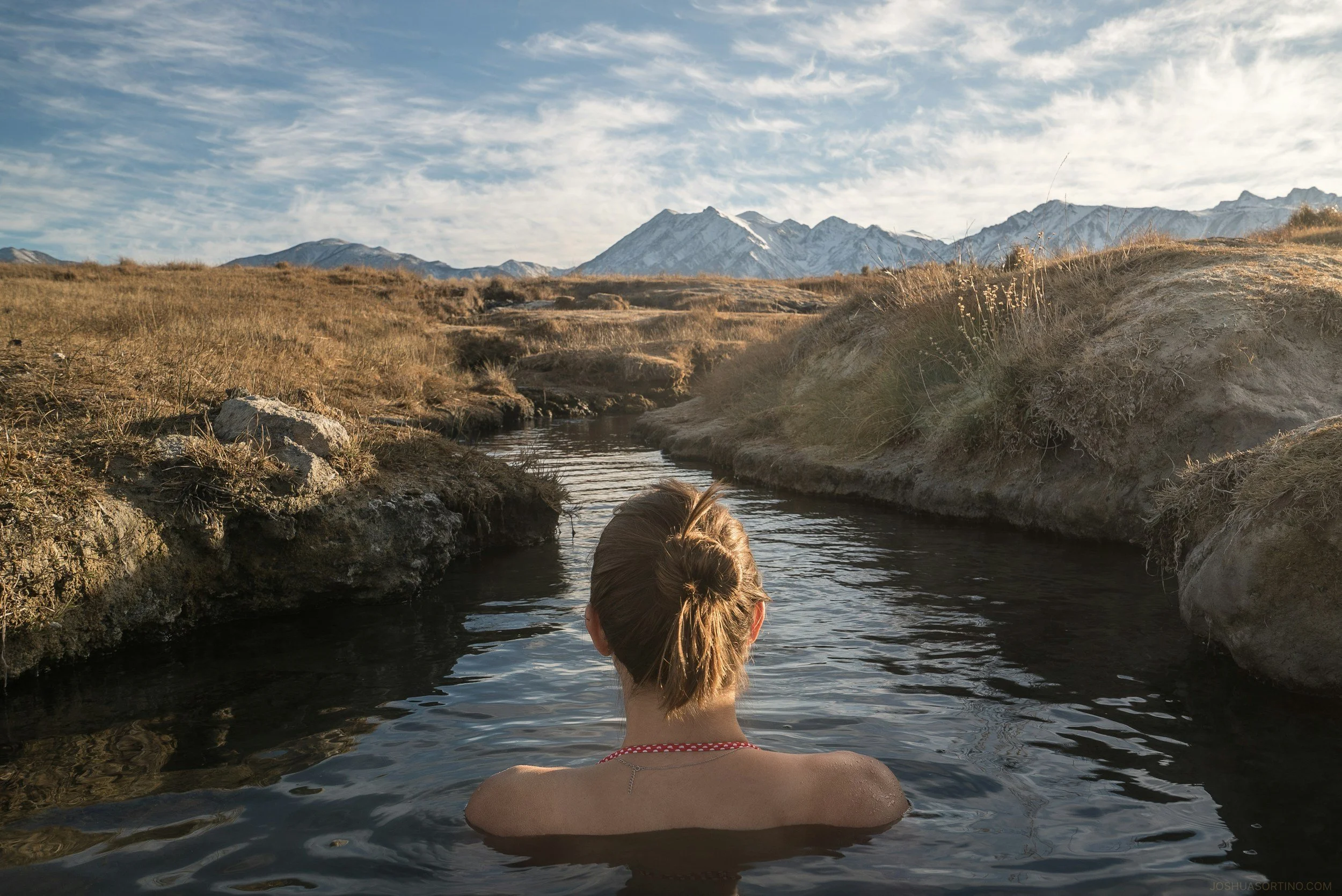 A woman with blond hair in a bun soaking in a natural hot spring surrounded by rocks and dry grass, with snow-capped mountains and a blue sky with clouds in the background.