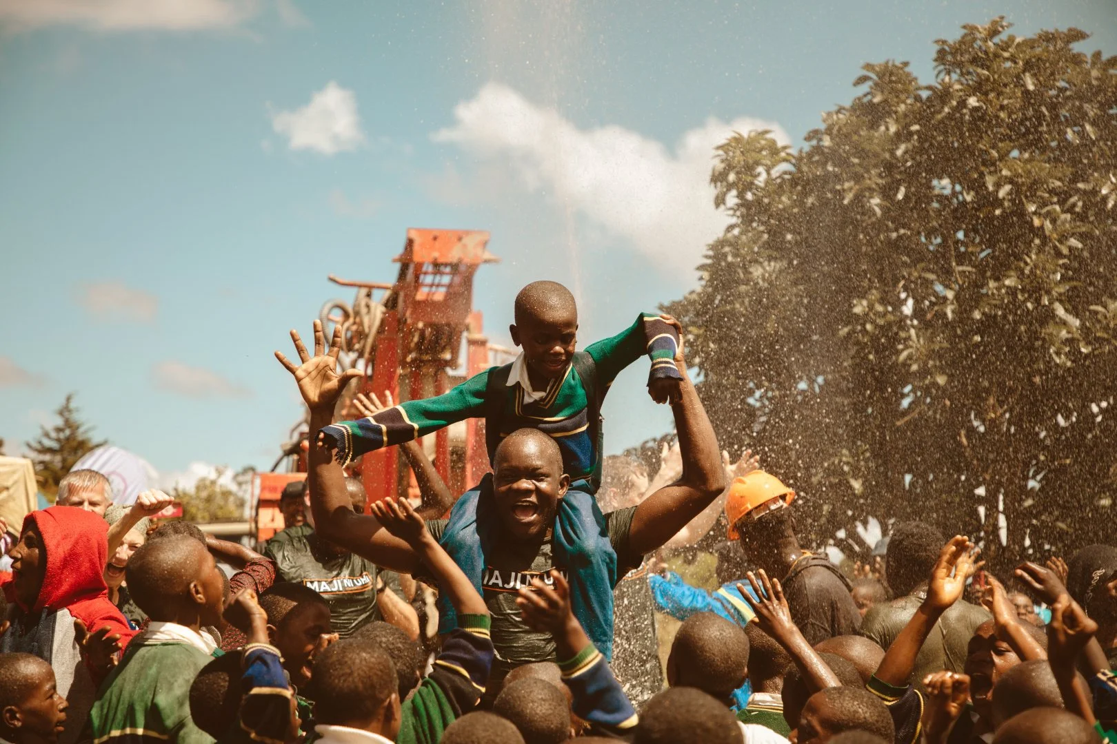 A joyful crowd of people, with one man lifting a young boy onto his shoulders in celebration, outdoors on a sunny day with trees and a water spray in the background.