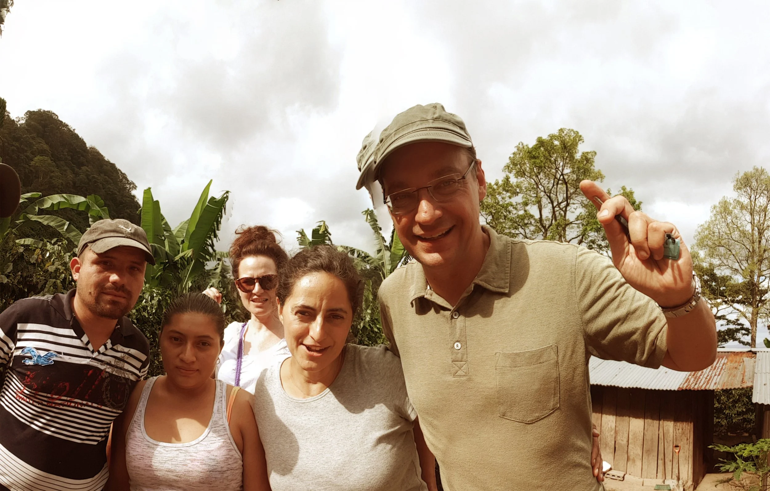 Group of five people outdoors in a rural setting, smiling and posing for a photo, with trees and a wooden structure in the background.