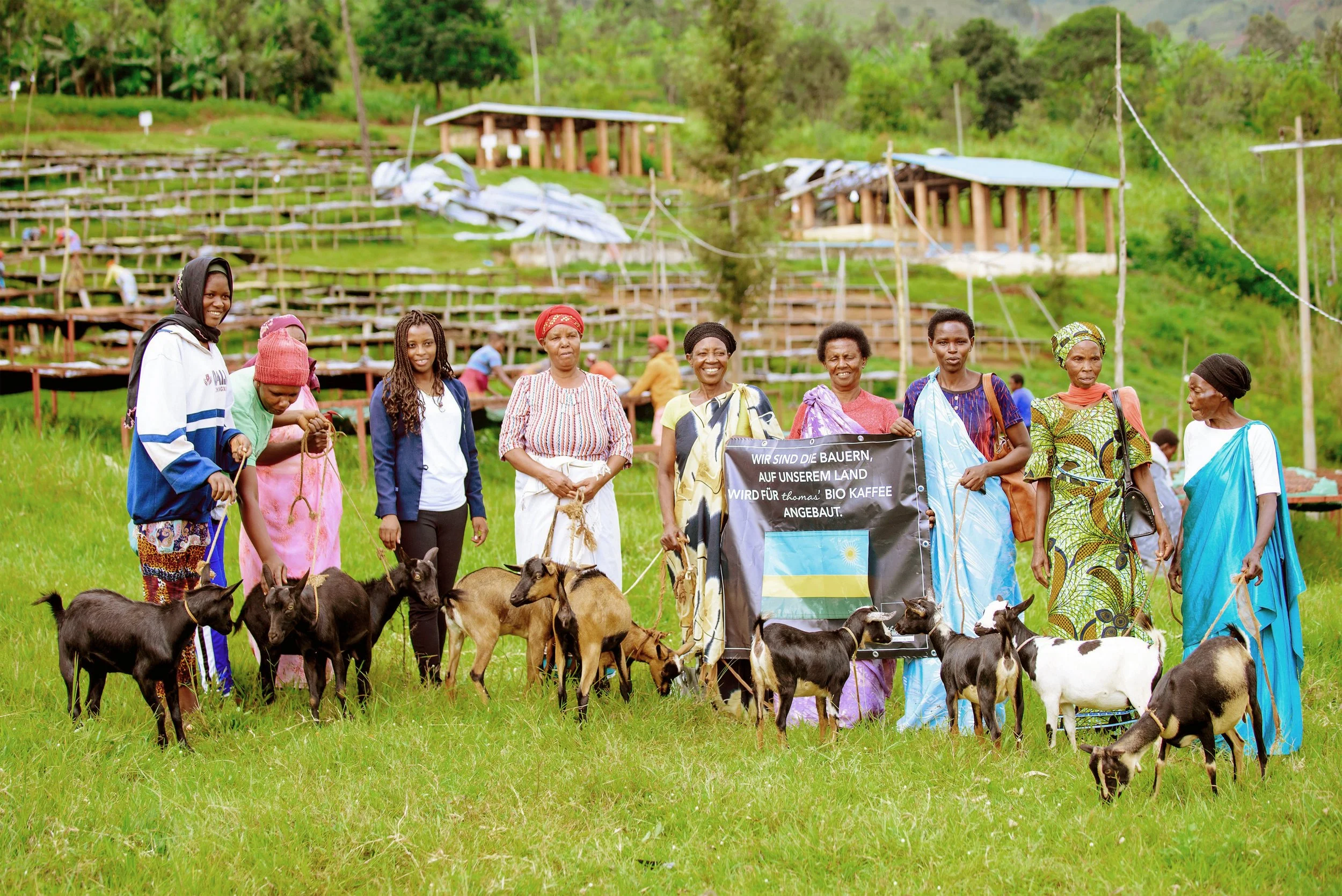 Group of women and girls standing in a grassy field with goats, holding a banner with a flag. The women are dressed in colorful traditional and modern clothing. The background shows a hillside with wooden structures and people working.