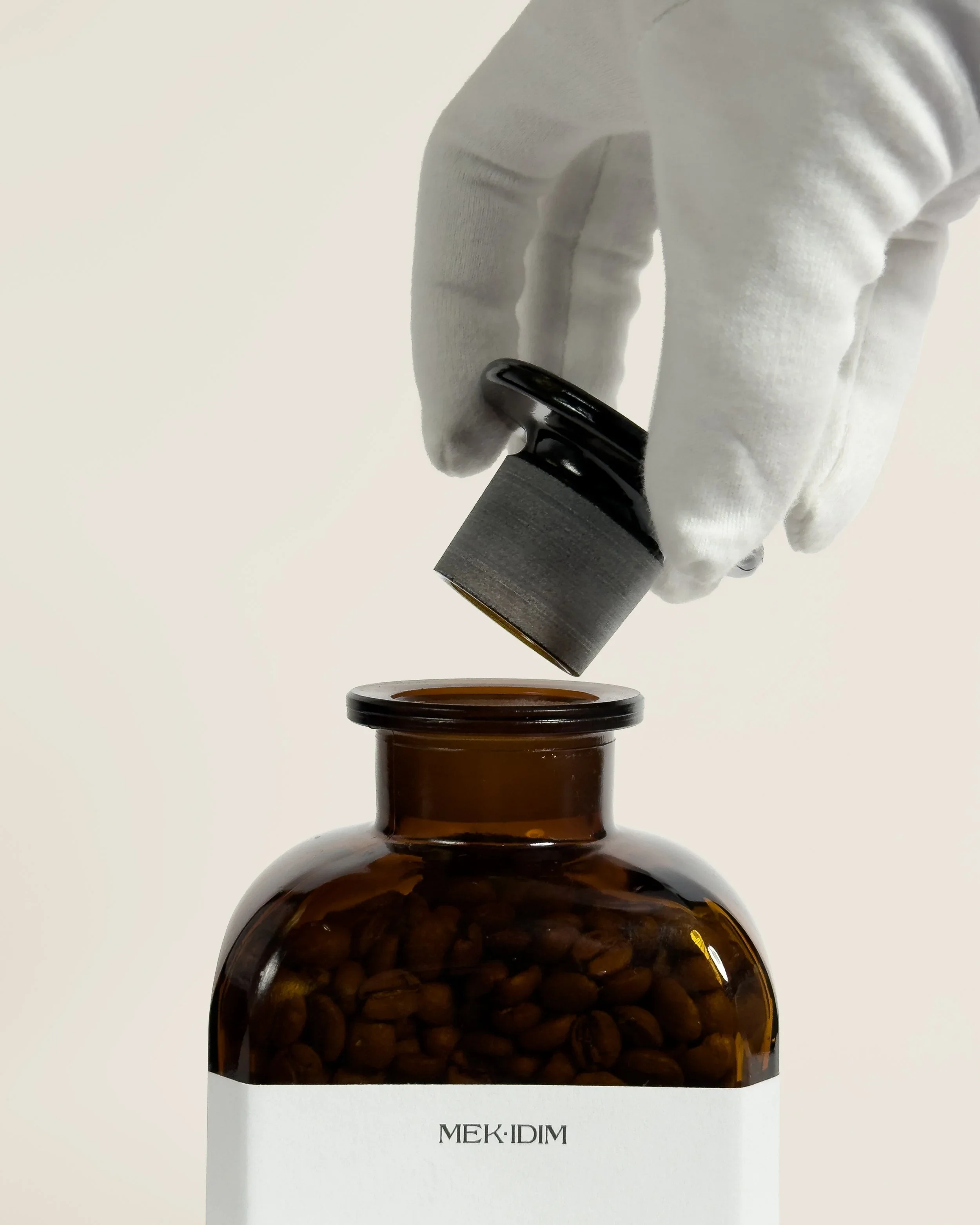 A gloved hand lifting a black capsule from a brown glass bottle labeled 'MEK IDIM' containing coffee beans.
