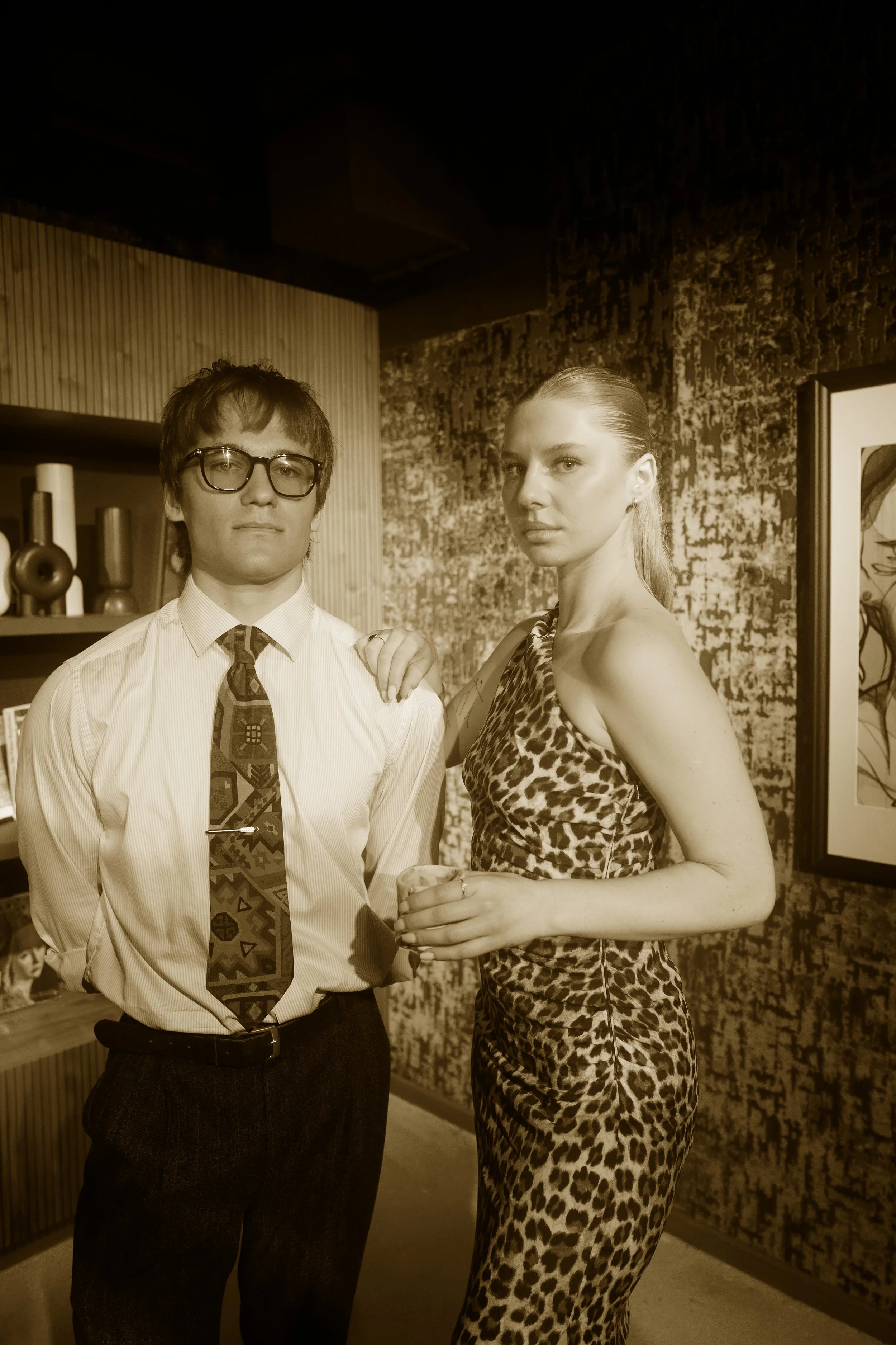 Two people standing together in an indoor setting, one man with glasses wearing a tie and a woman in a leopard print dress, with decorative objects and artwork in the background.