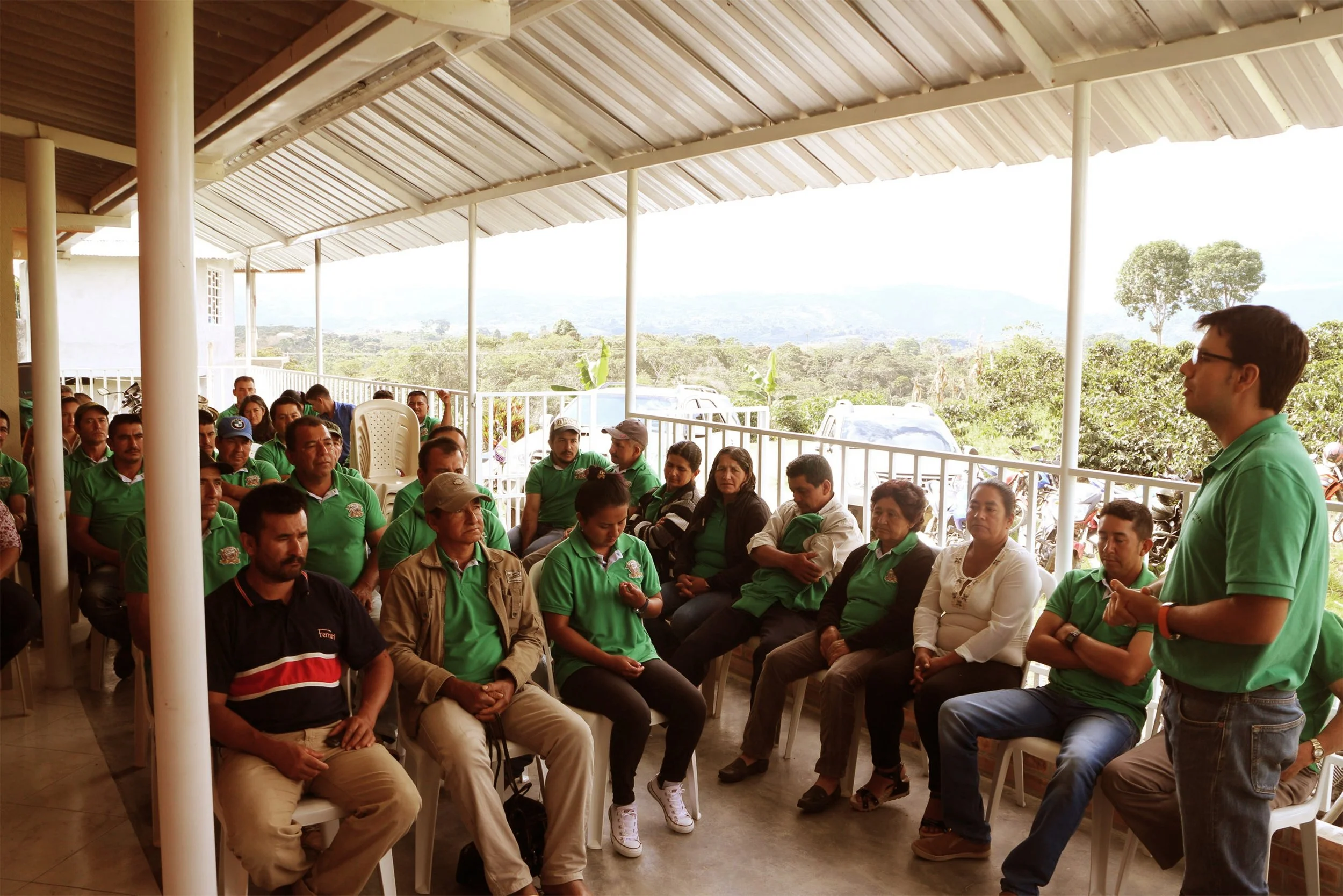 A man in a green polo shirt giving a presentation to a group of people seated on chairs in an outdoor covered area. The group is listening attentively, and the background shows a scenic landscape with trees and hills.