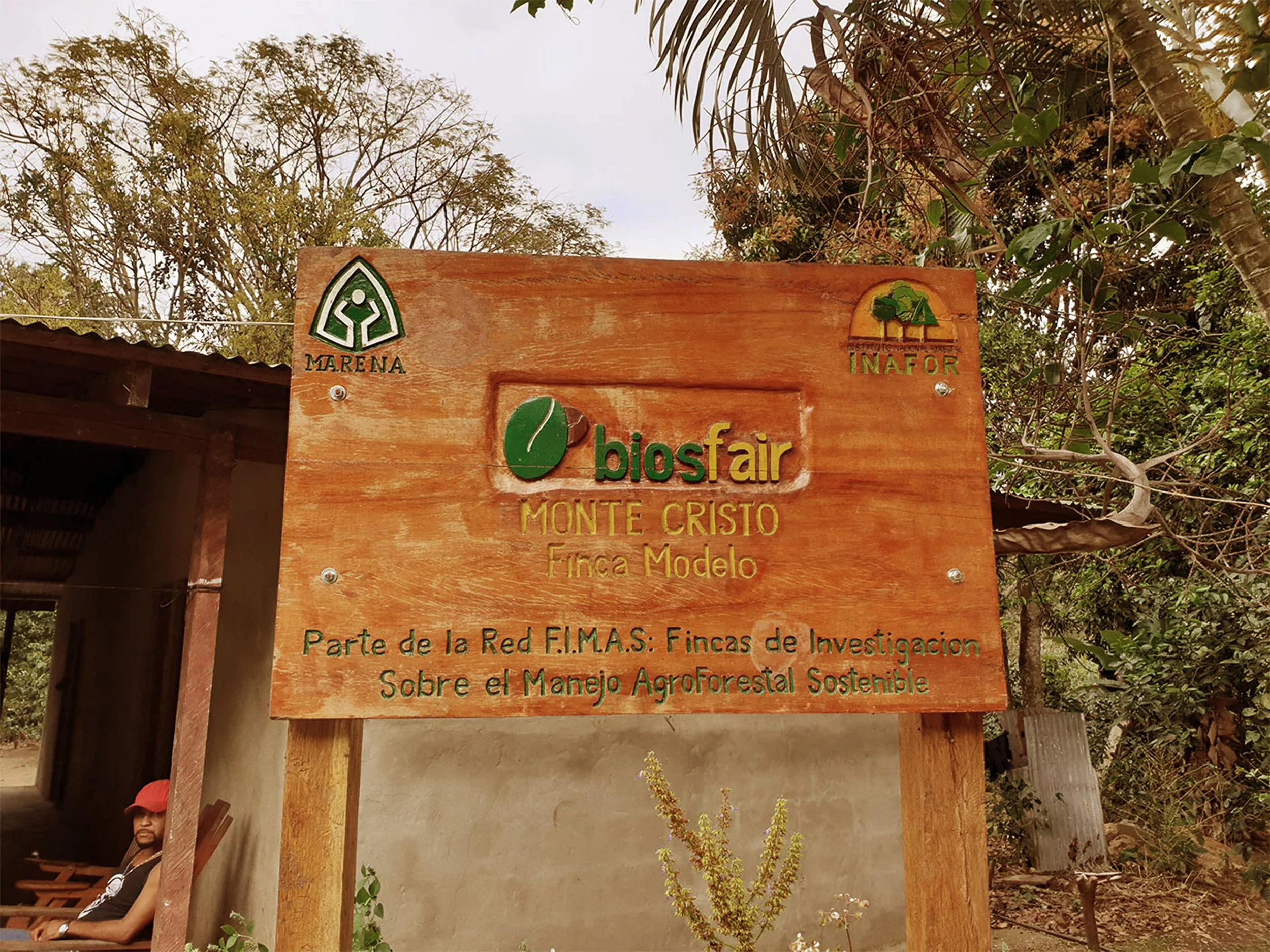 Wooden sign with logos and text in Spanish, indicating a biosafety fair at Monte Cristo Finca Modelo, part of the F.I.M.A.S. network about sustainable agroforestry management. The background shows trees and part of a structure with a man sitting.
