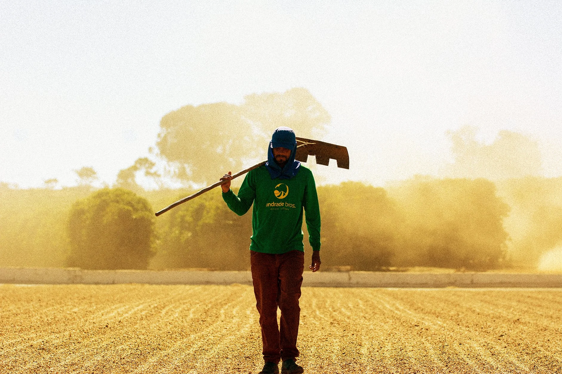 A person walking across a dirt field with a long-handled tool on their shoulder, wearing a green shirt and a face covering, with trees and a clear sky in the background.