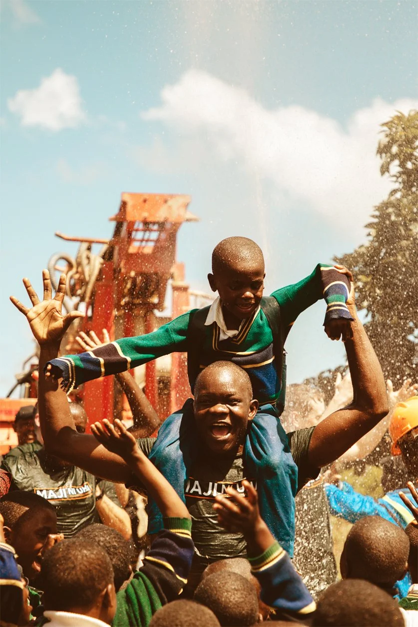 A group of children outdoors, with a joyful African boy sitting on an adult's shoulders, raising his hand. Others are cheering around them, and there is a playground structure in the background under a partly cloudy sky.