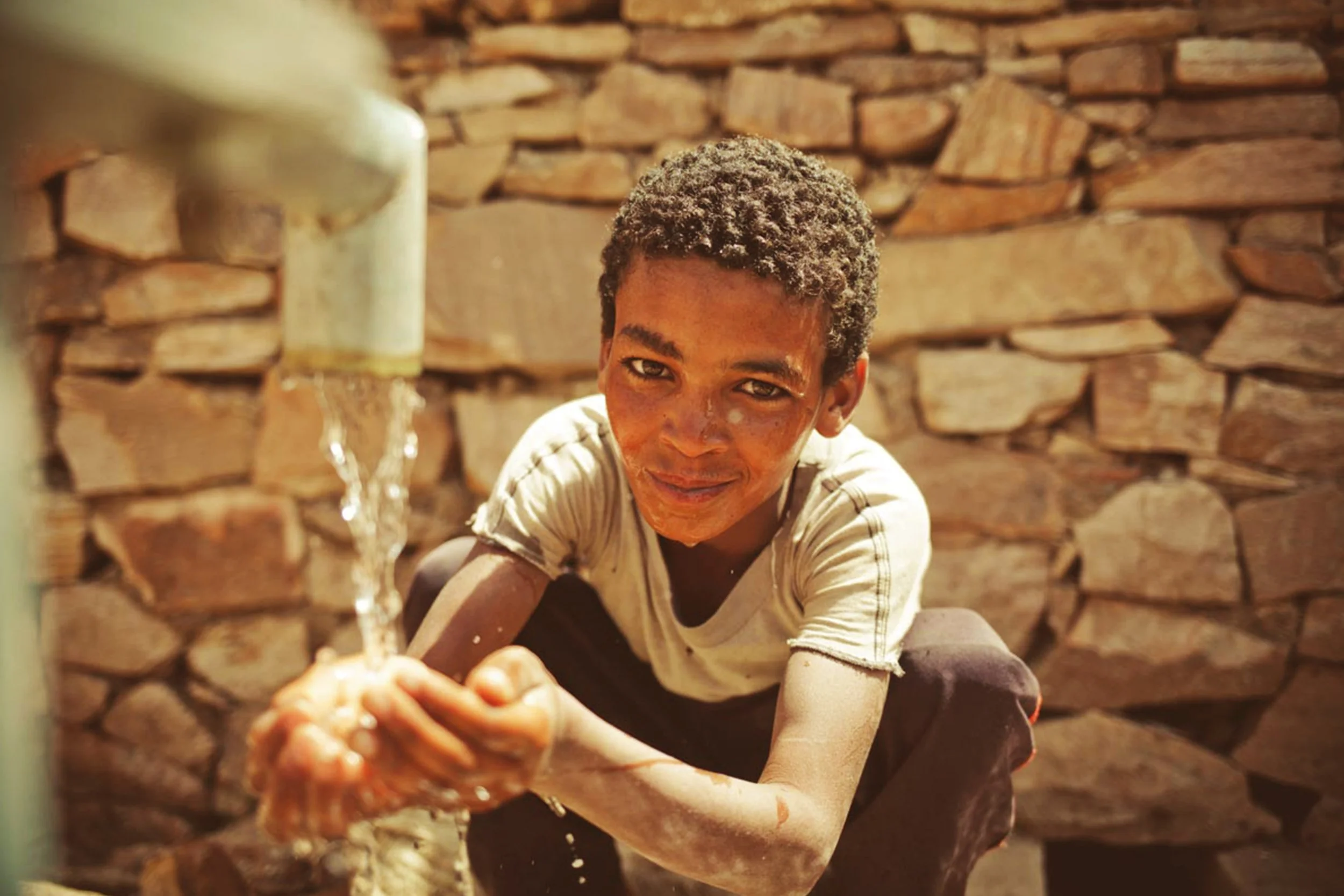 A young boy squatting and smiling while reaching out for water from a faucet, with a stone wall in the background.