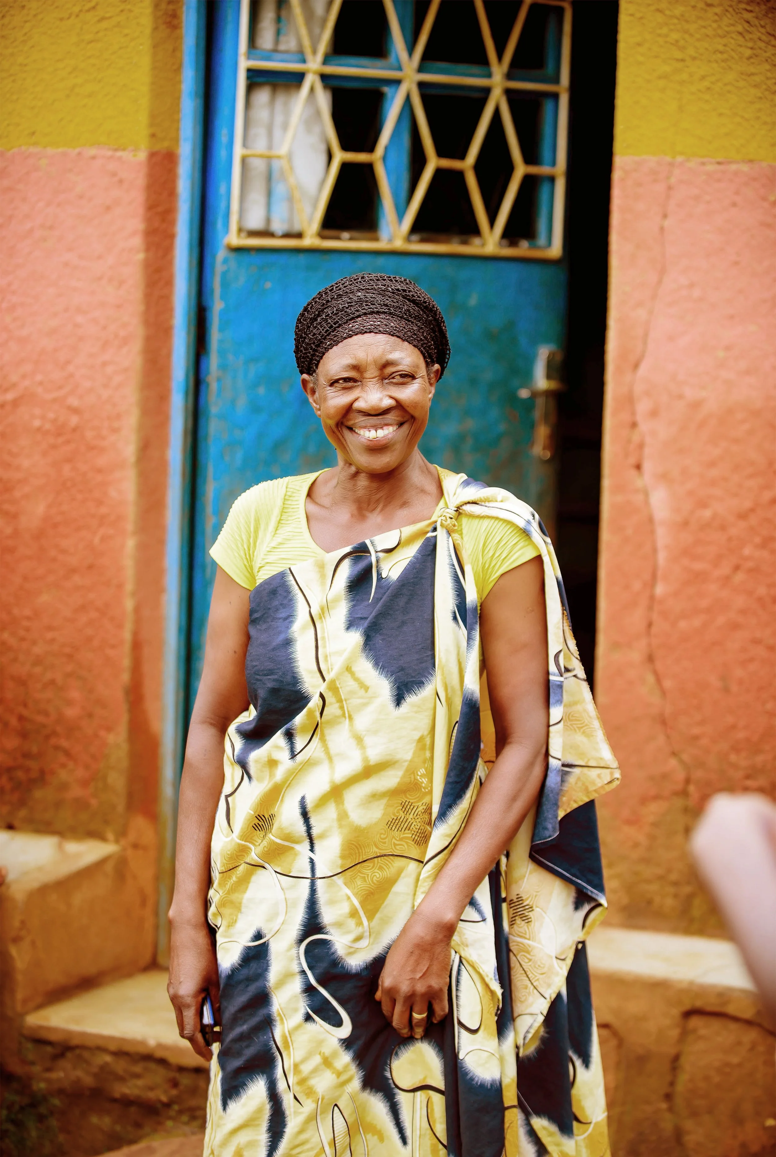 A smiling woman wearing a vibrant yellow and black patterned dress with a matching shawl, standing in front of a blue door with yellow and black window bars, against a multicolored wall in an outdoor setting.