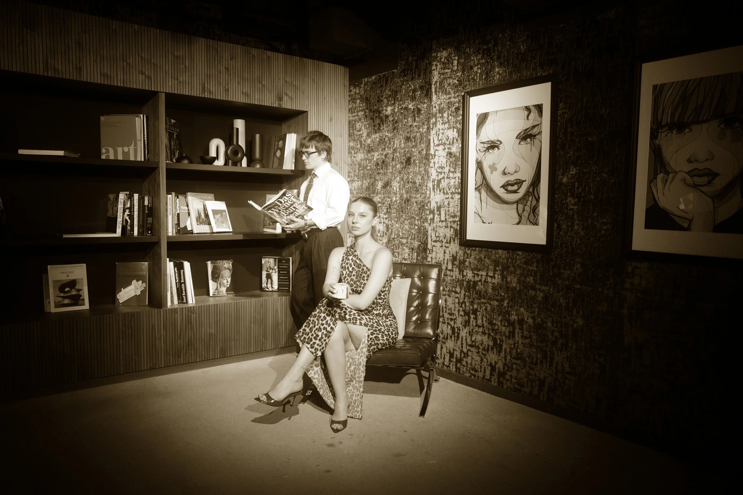 A black and white photo of a woman sitting on a chair in an art gallery, wearing a leopard print dress and heels, holding a mug. A man in glasses and formal attire stands nearby, reading a book, with a bookshelf and framed artwork on the wall behind them.