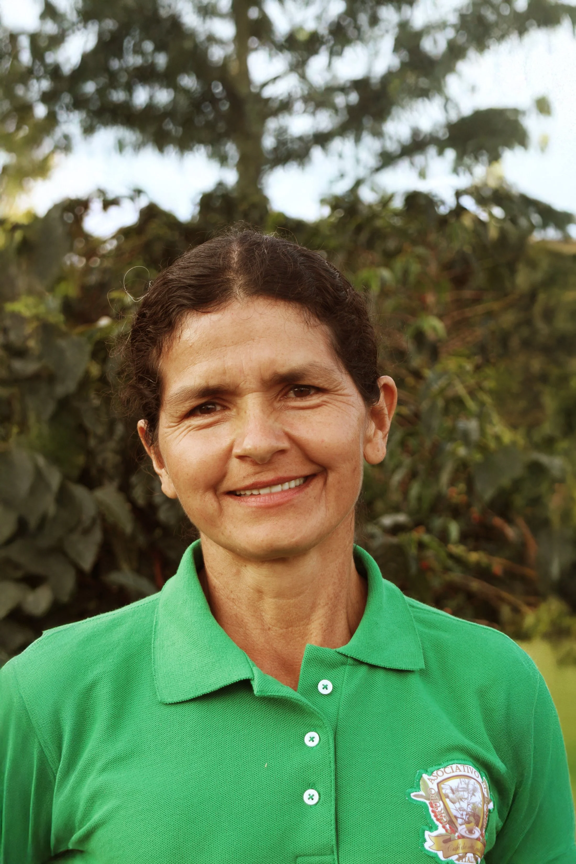 Portrait of a woman wearing a green polo shirt with a logo, smiling outdoors with trees in the background.