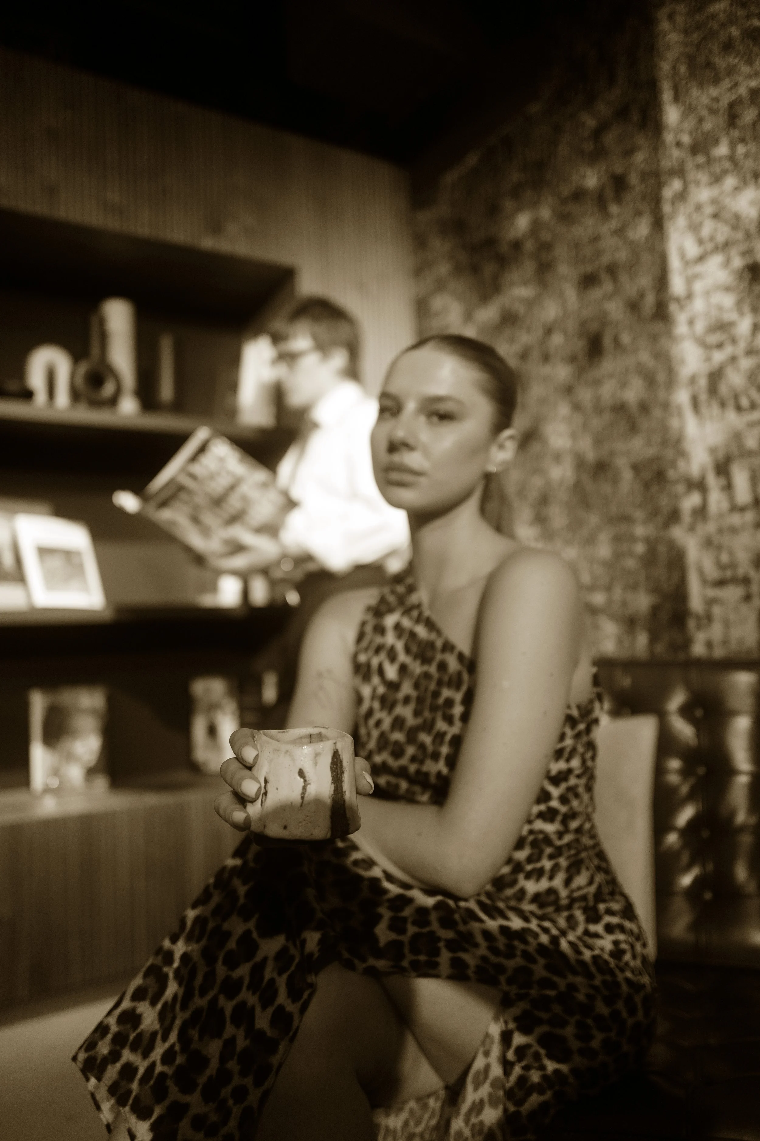A woman with a leopard print dress sitting on a chair holding a mug, with a person reading a book in the background in a cozy room.