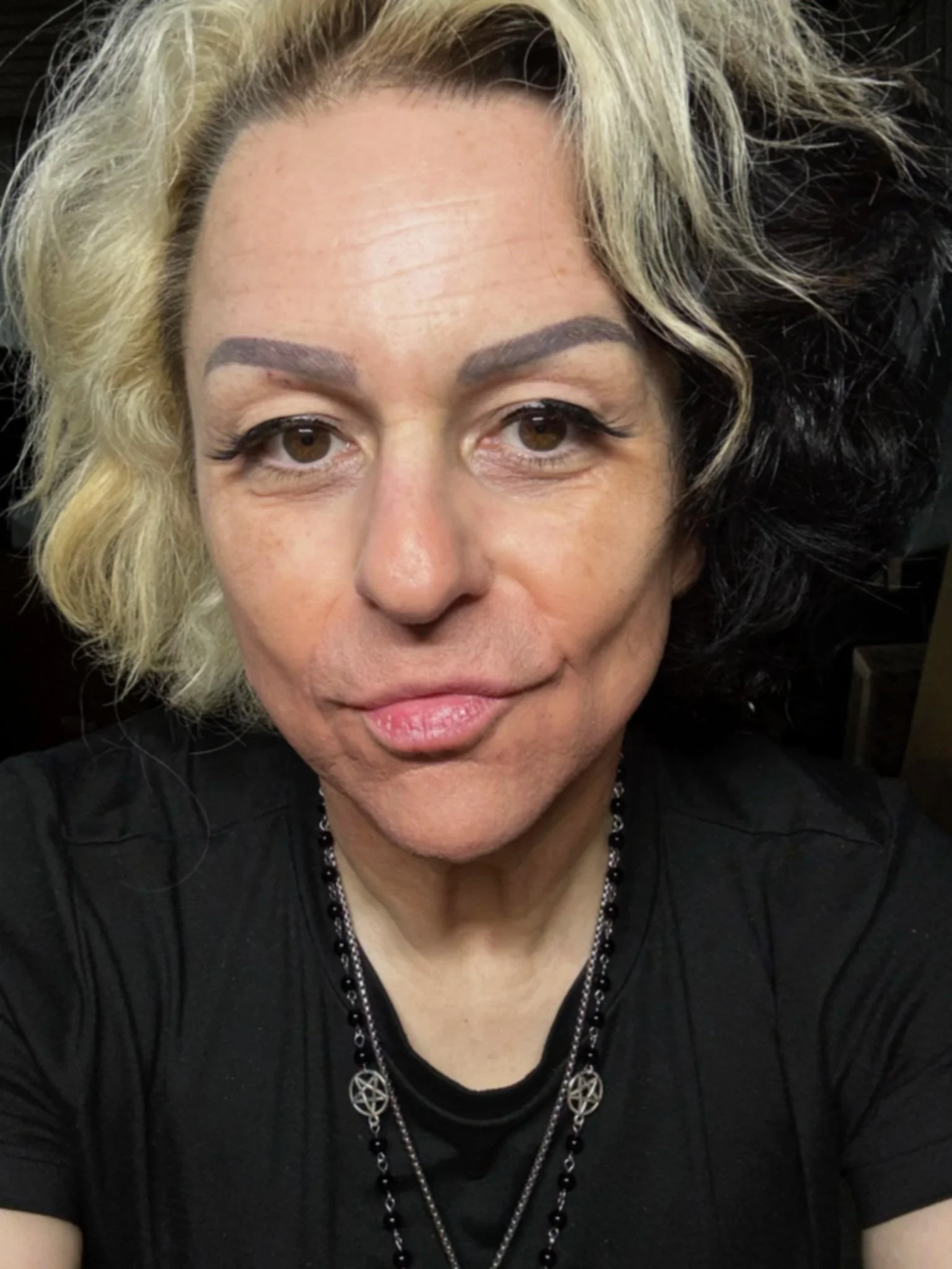 A woman with blonde and black hair, wearing dark makeup, jewelry, and a black top.