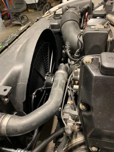 Close-up of a BMW engine bay with Koop's Kustoms electric fan kit, hoses, radiator, and engine components.