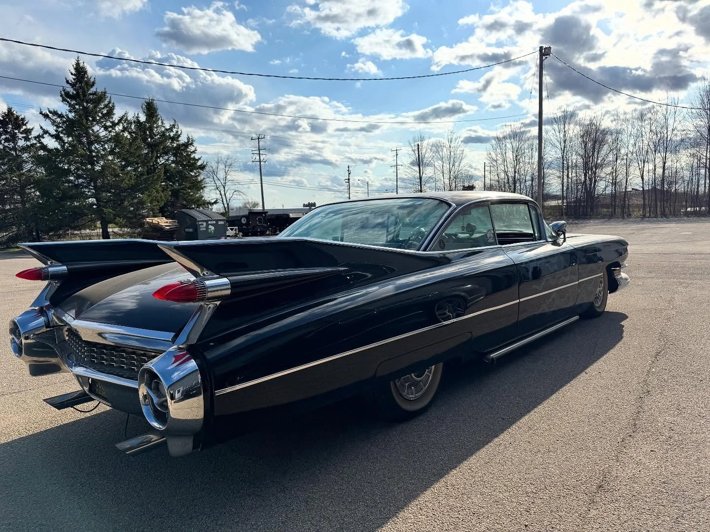 A vintage black 1959 Cadillac car parked on a paved lot, with a cloudy sky and leafless trees in the background.