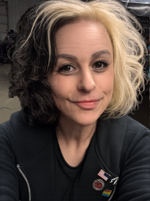 A woman with half black and half blonde curly hair taking a selfie, smiling slightly, in a room with dark background, wearing a dark top with pins including an American flag.