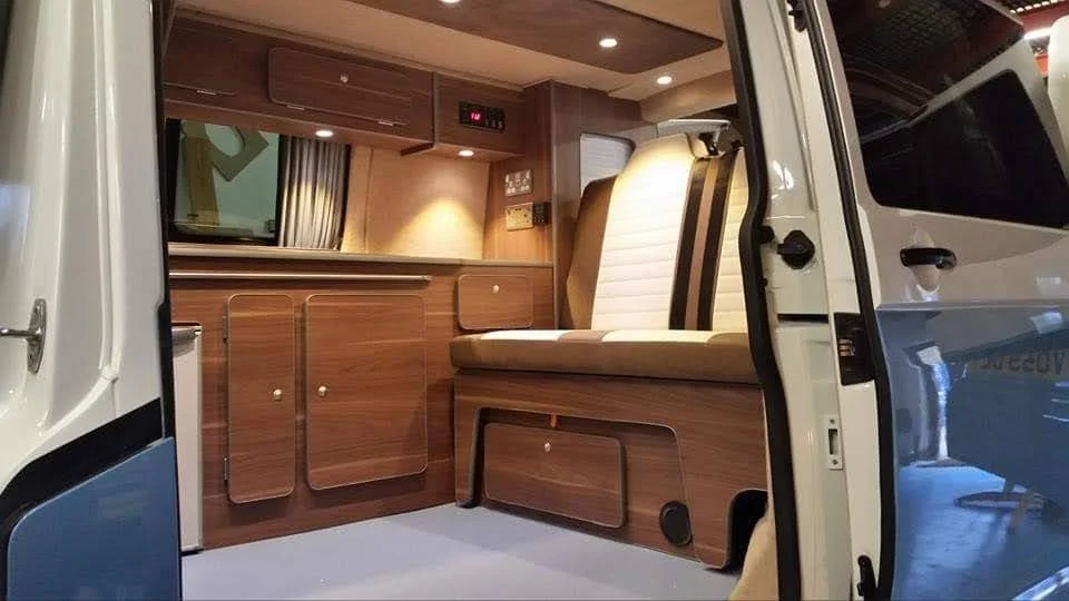 Interior of a camper van with wooden cabinetry, a window with curtains, and a cushioned bench seat.