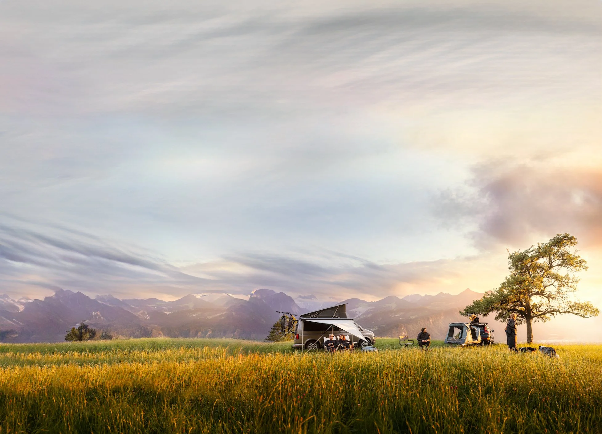 People camping and relaxing in a grassy field with tents and a van, mountains in the background, during sunset.