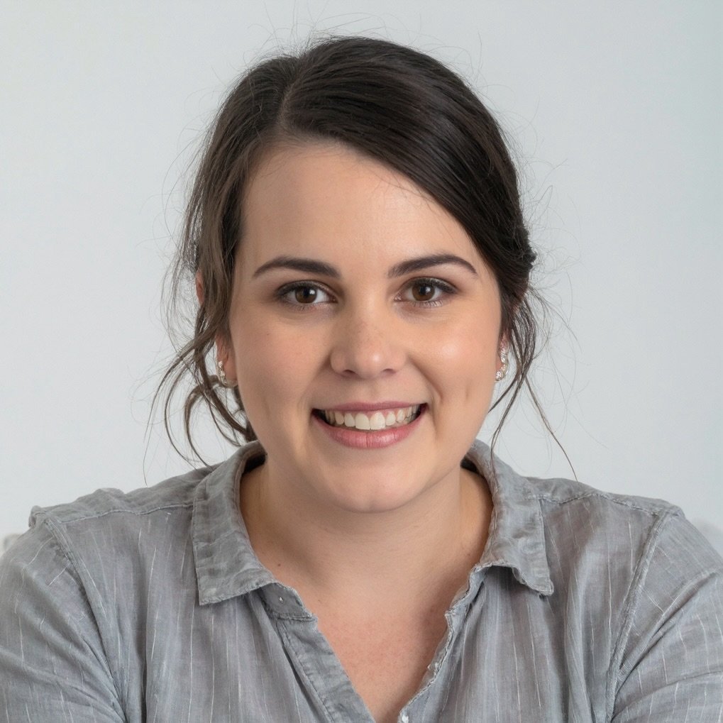Close-up portrait of a woman with dark brown hair, brown eyes, and a light complexion, smiling, wearing a gray collared shirt, against a plain light background.