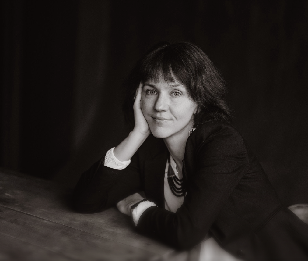 A woman writer sits at a table in a blazer, looking at the camera, her face resting on her hand. Black and white
