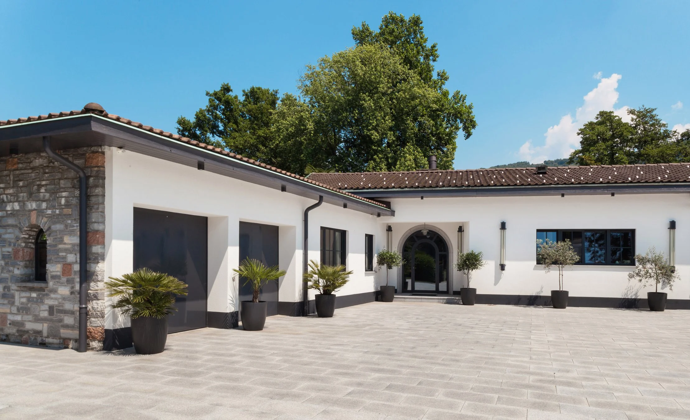 Casa moderna con pareti bianche, finestre nere, porta d'ingresso arcuata, piante in vasi neri e un cortile pavimentato, con alcuni alberi verdi sullo sfondo e un cielo azzurro.