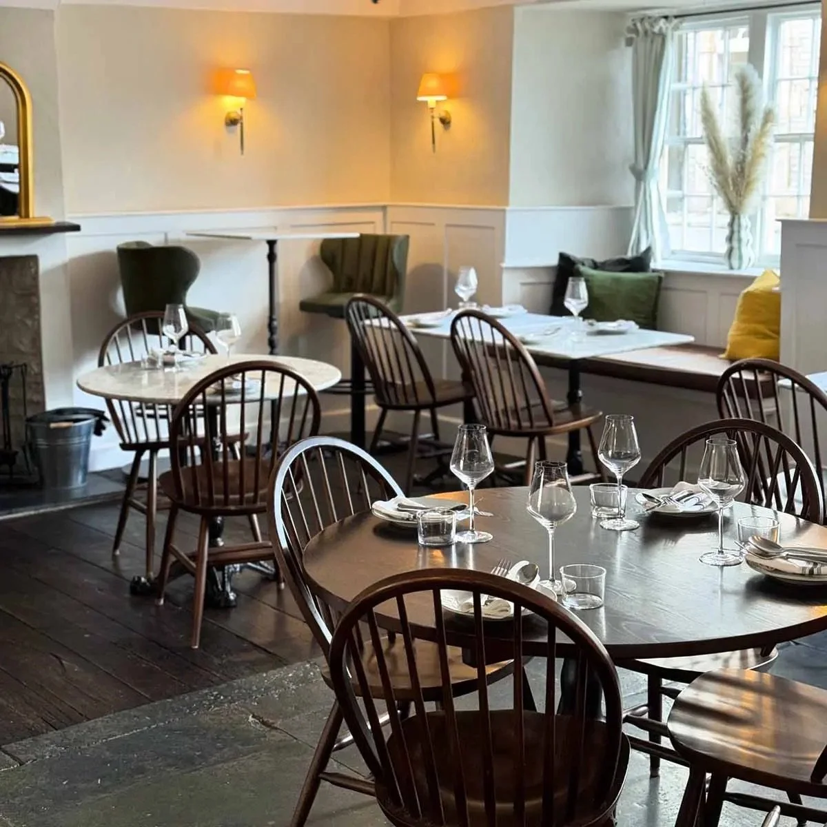 A cozy restaurant interior with wooden tables set with wine glasses, water glasses, plates, and silverware. There are wooden and upholstered chairs, a window seat with pillows, and warm wall-mounted lighting.