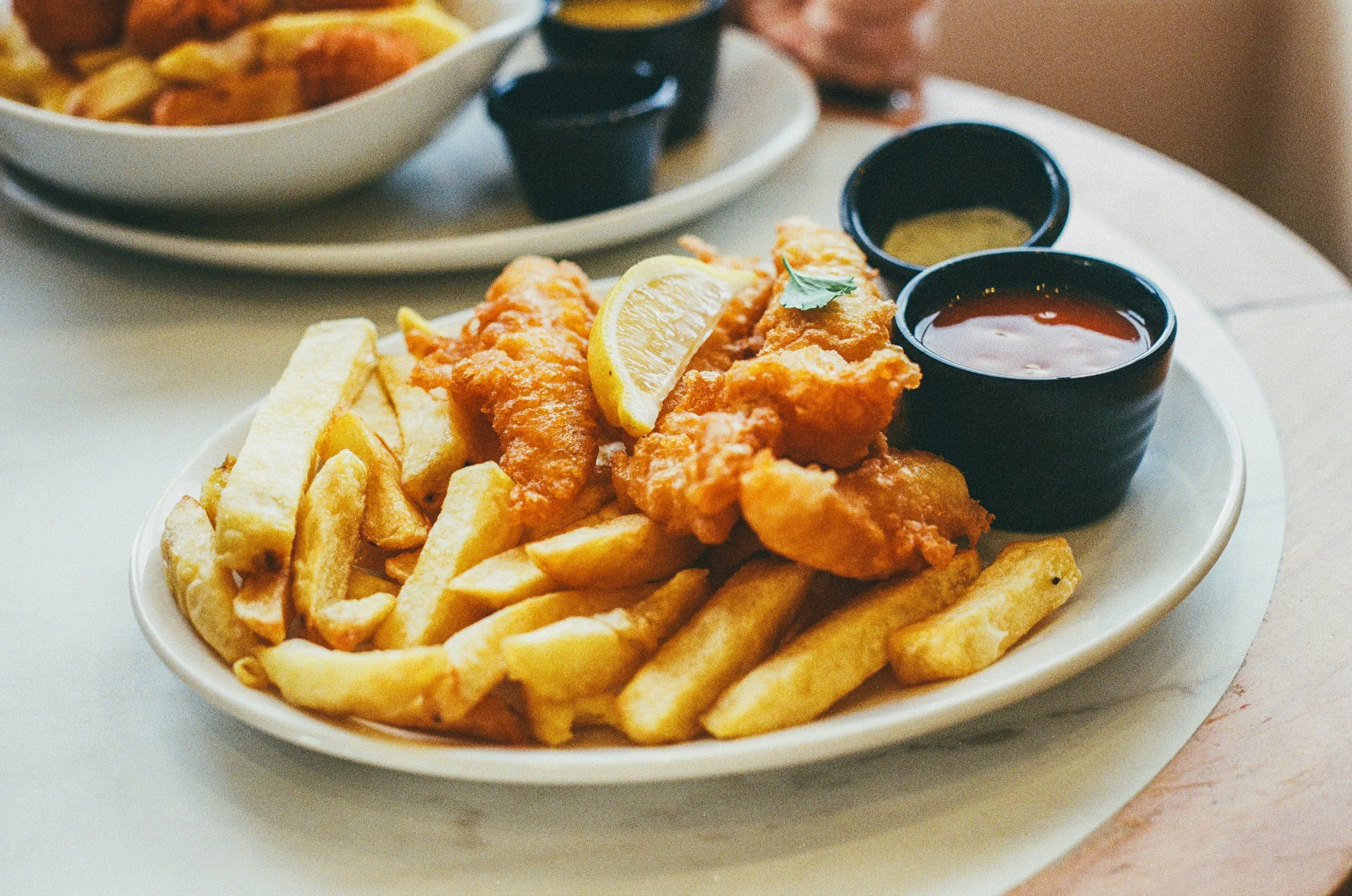 A plate of French fries topped with fried fish fillets, garnished with lemon wedges and parsley, served with two small bowls of dipping sauces, one red and one yellow.