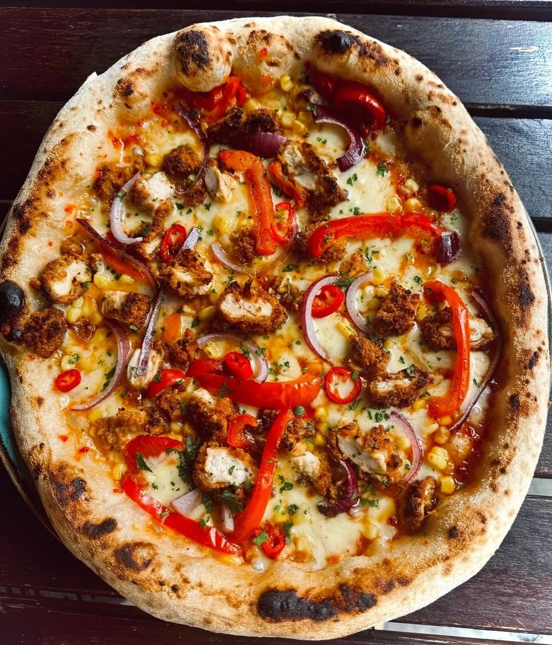 A pizza topped with red bell peppers, red onions, corn, and pieces of fried chicken on a dark wooden table.