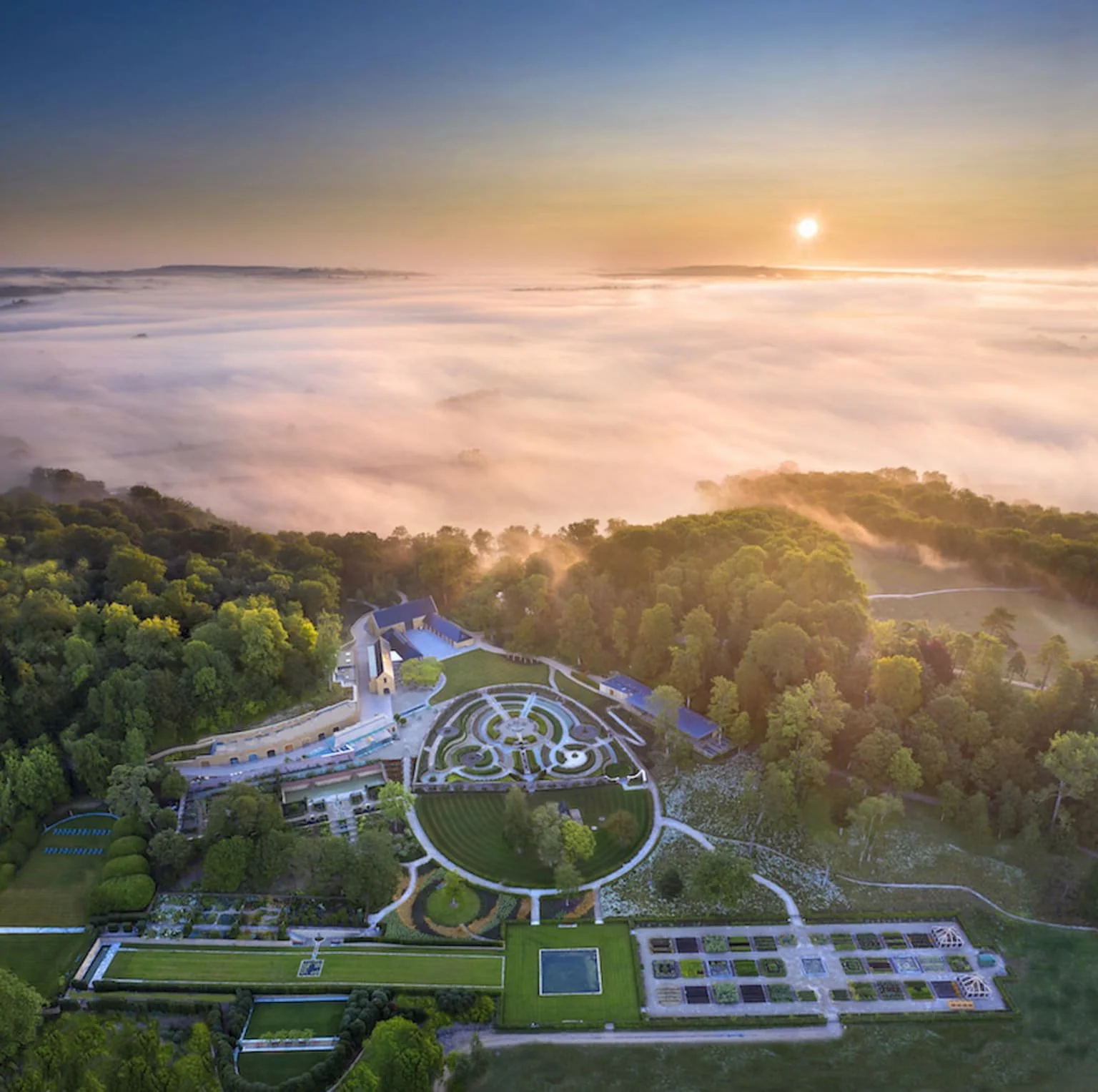 Aerial view of a botanical garden with neatly arranged flower beds and pathways surrounded by lush green trees, with a fog-covered landscape and sunrise in the background.