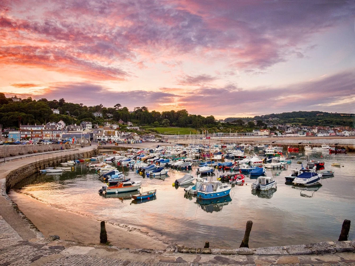 Harbor full of boats at sunset with colorful sky and buildings in the background.