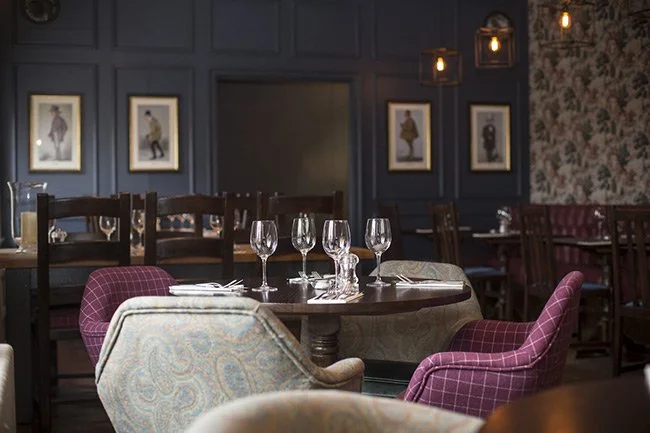 A restaurant interior with a round table set with four wine glasses, surrounded by upholstered chairs, dark blue paneled walls decorated with framed artwork and wall sconces.