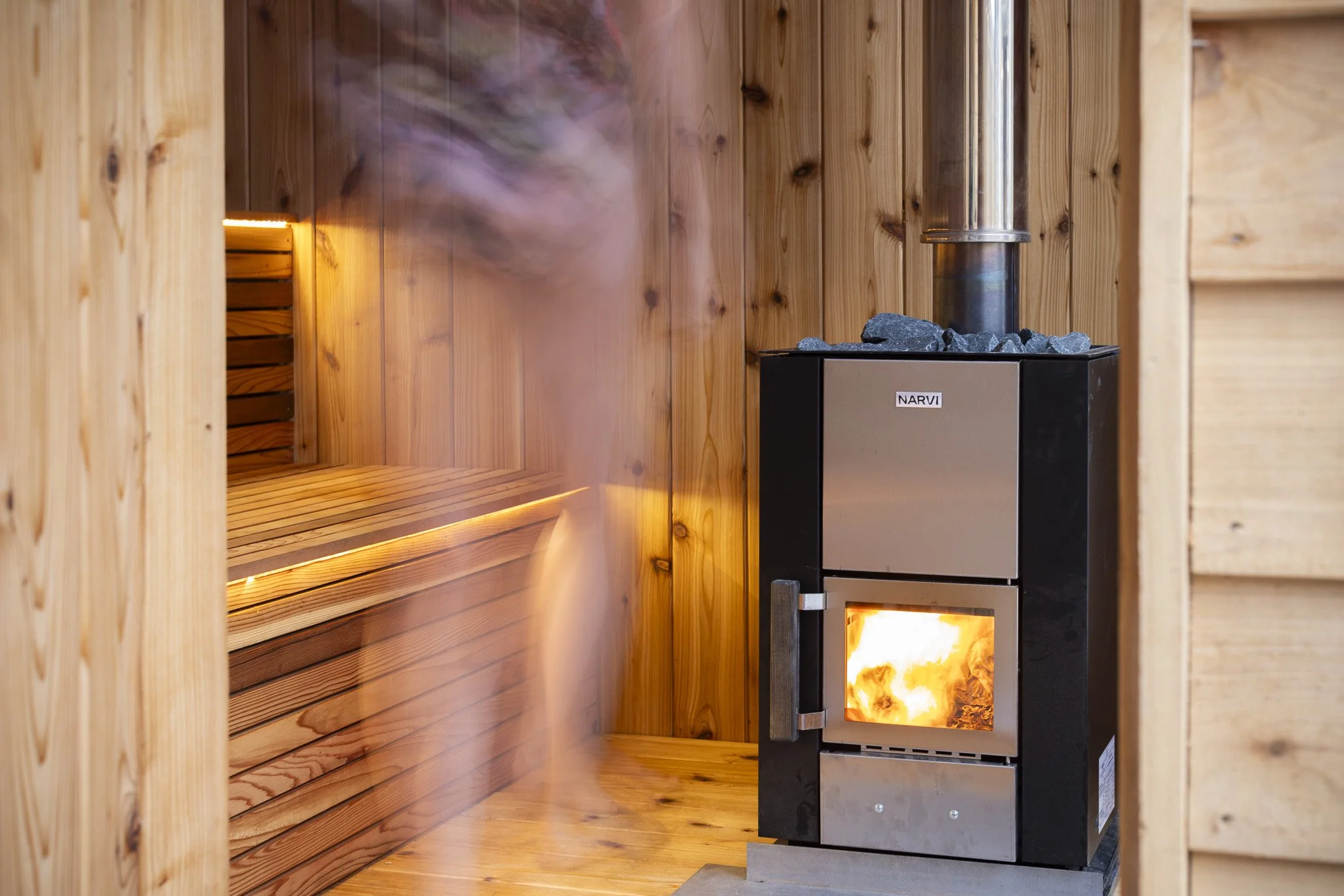 Wood-paneled sauna room with a black heater emitting a fire inside, and a wooden bench on the left side of the room.