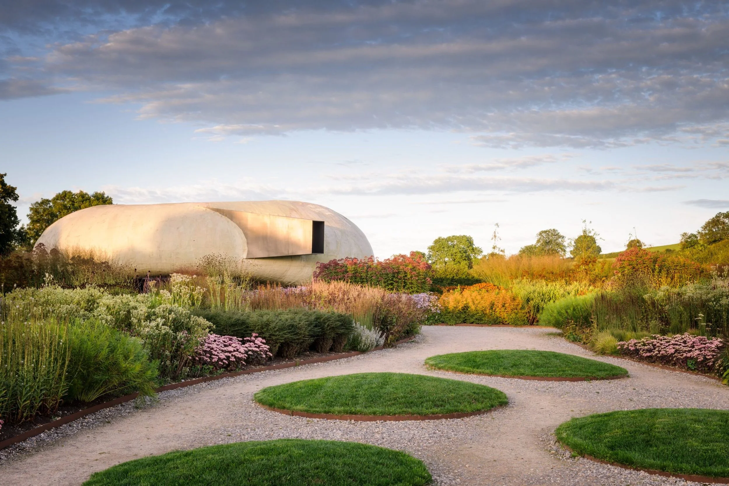 Modern oval-shaped building made of concrete in a garden with colorful flowers and a gravel pathway