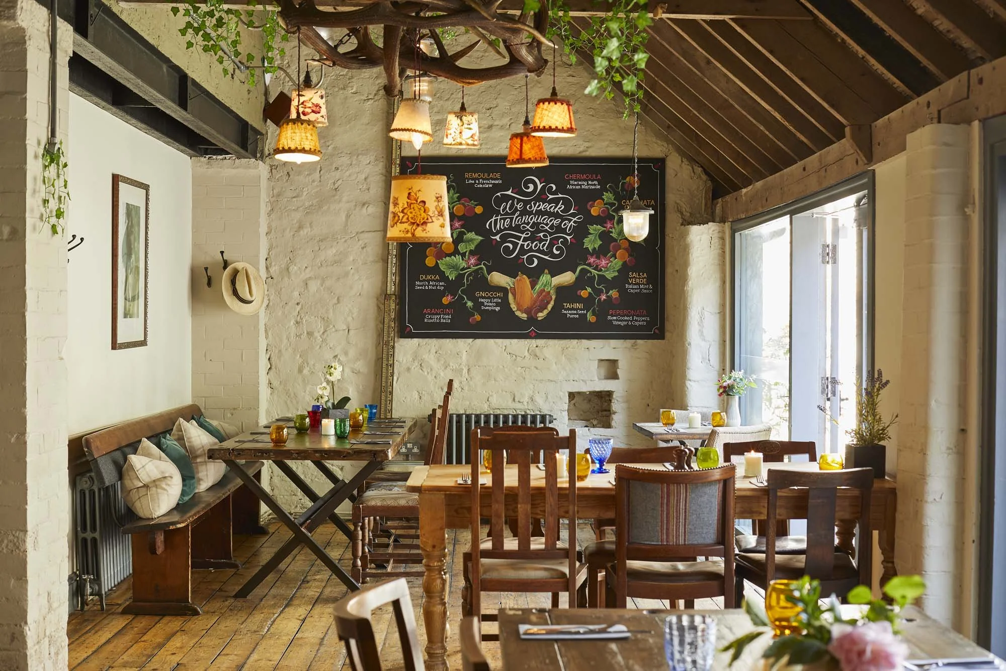 Cozy restaurant interior with wooden furniture, colorful glassware, and a large blackboard with food descriptions, decorated with hanging lamps, plants, and paintings.