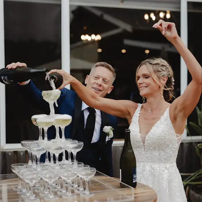 Bride and groom celebrating, pouring champagne into a pyramid of glasses at their wedding reception.