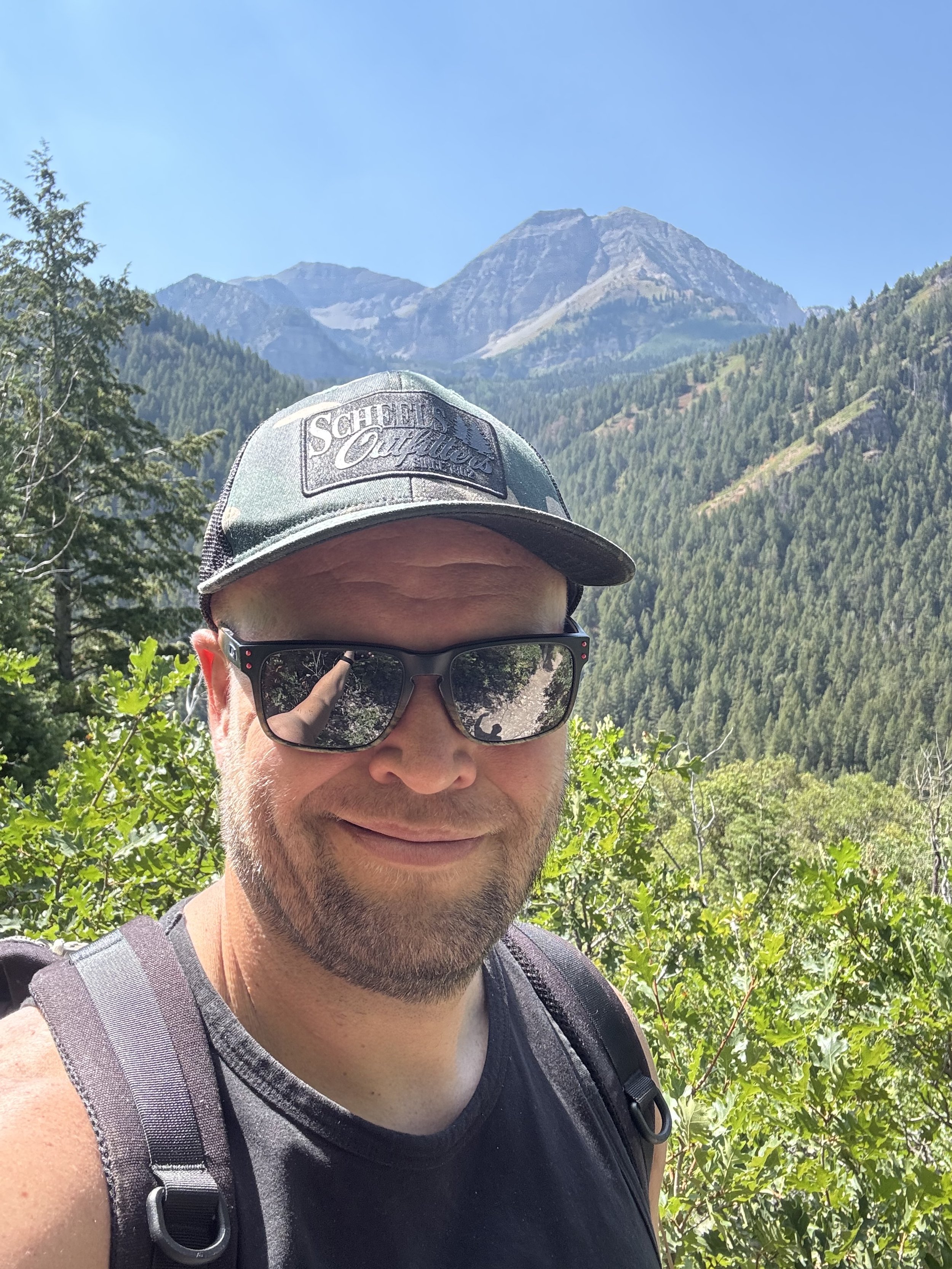 A man wearing sunglasses and a baseball cap, smiling outdoors among green foliage with mountains and a clear blue sky in the background.