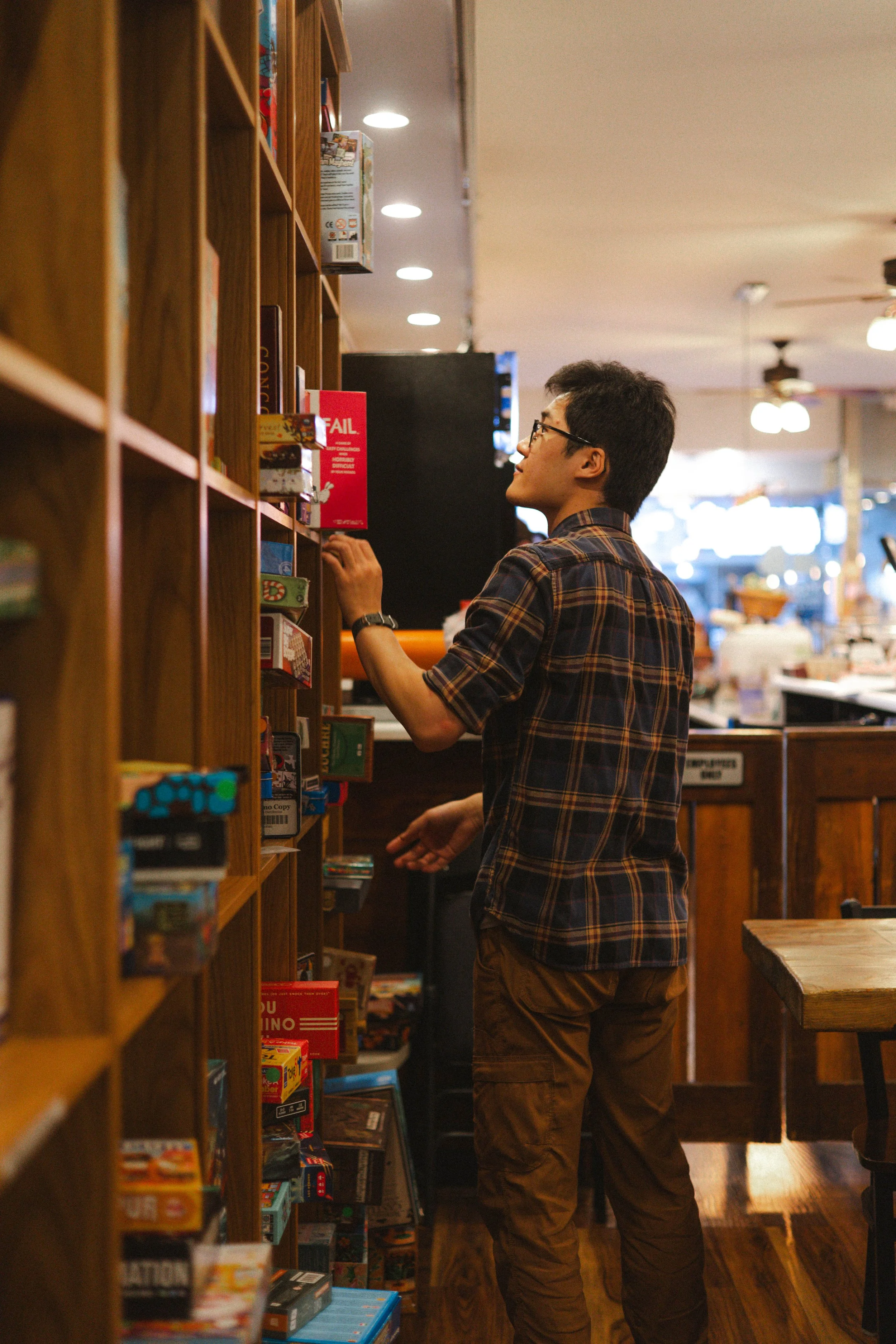 Young man with glasses browsing board games on a wooden shelf inside a store.