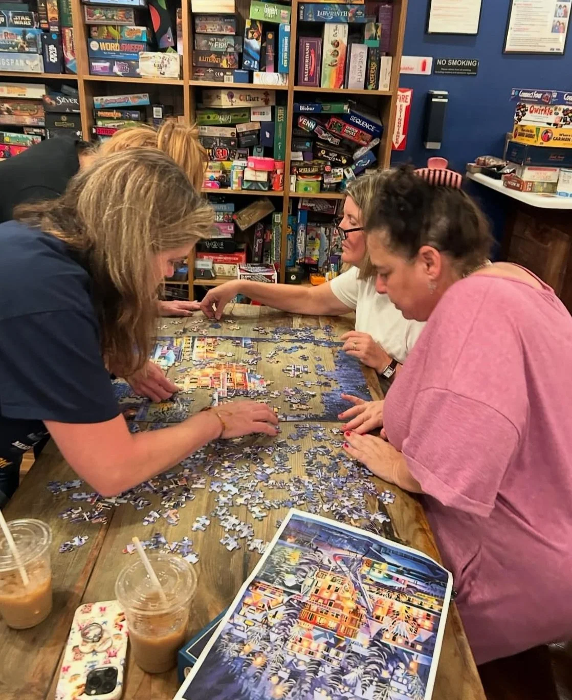 Four women working on assembling a colorful jigsaw puzzle at a wooden table in the cafe, with shelves filled with board games and puzzles in the background. Drinks are on the table.