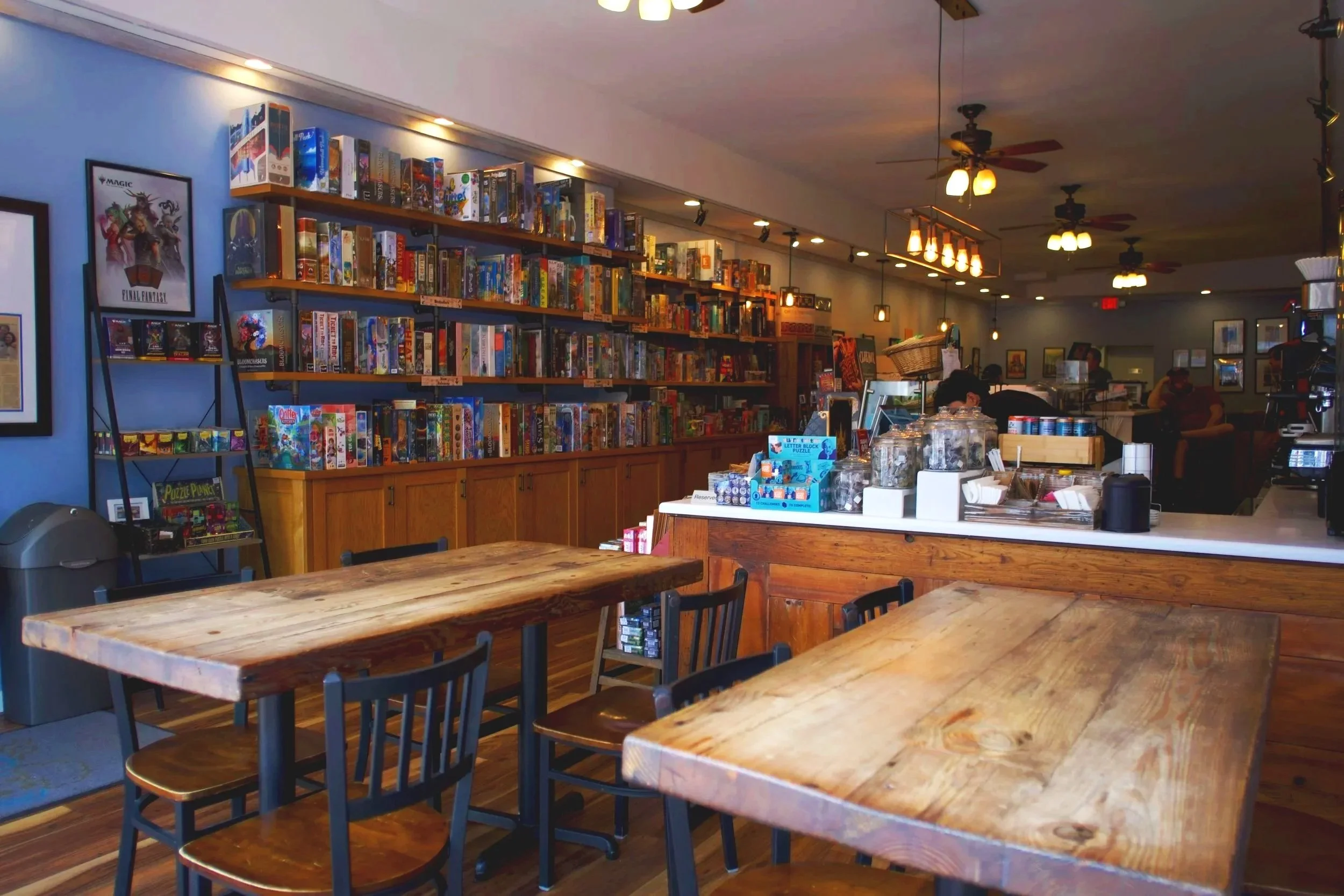 Interior of a cozy cafe or game store with wooden tables, black chairs, and shelves filled with board games and puzzles. The space has warm lighting, ceiling fans, and a counter area with jars and snacks in the background.