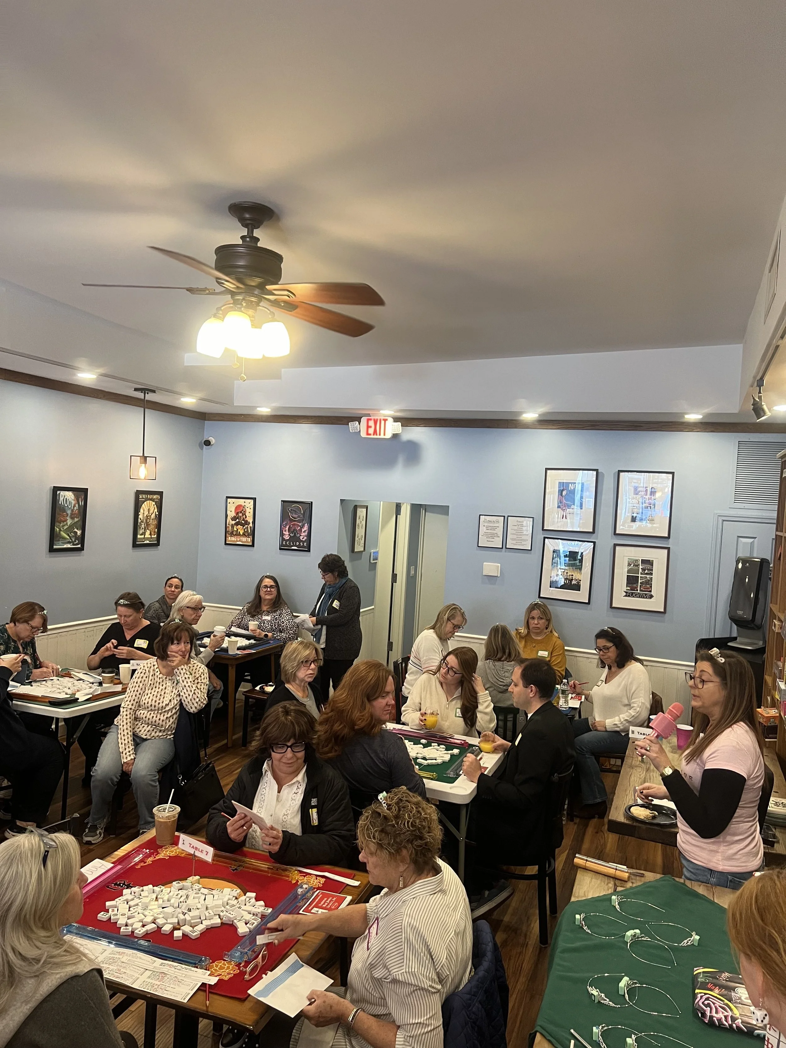 Group of women gathered in a room playing Mahjong, with some seated around tables and others standing. There are various framed pictures on the walls, and a ceiling fan with lights is overhead. The room has a casual, social atmosphere.