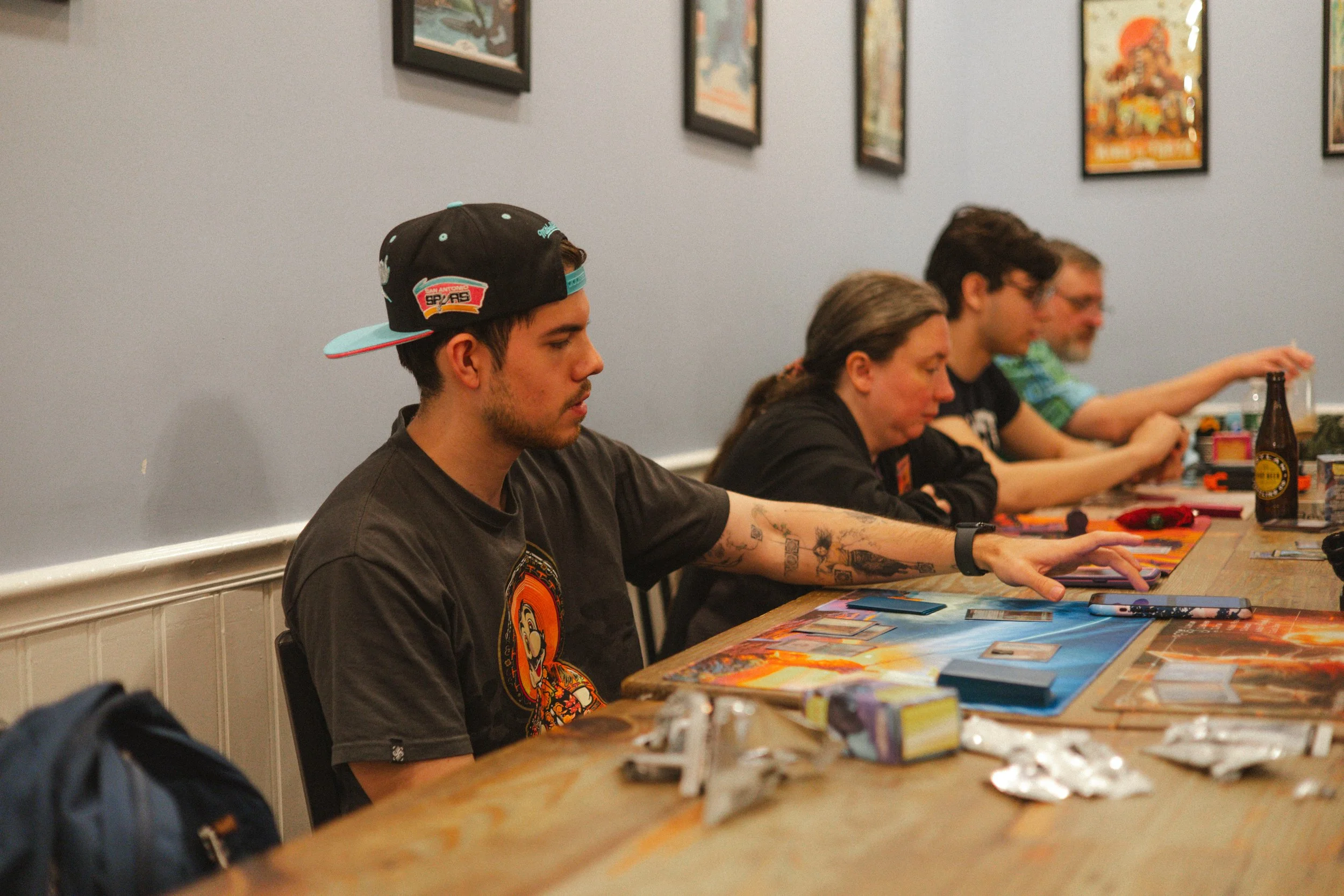 Four people are seated at a long table playing a card game indoors with framed artwork on the wall behind them. The person closest to the camera is a young man wearing a black cap backwards and a black T-shirt with a graphic design, reaching for his