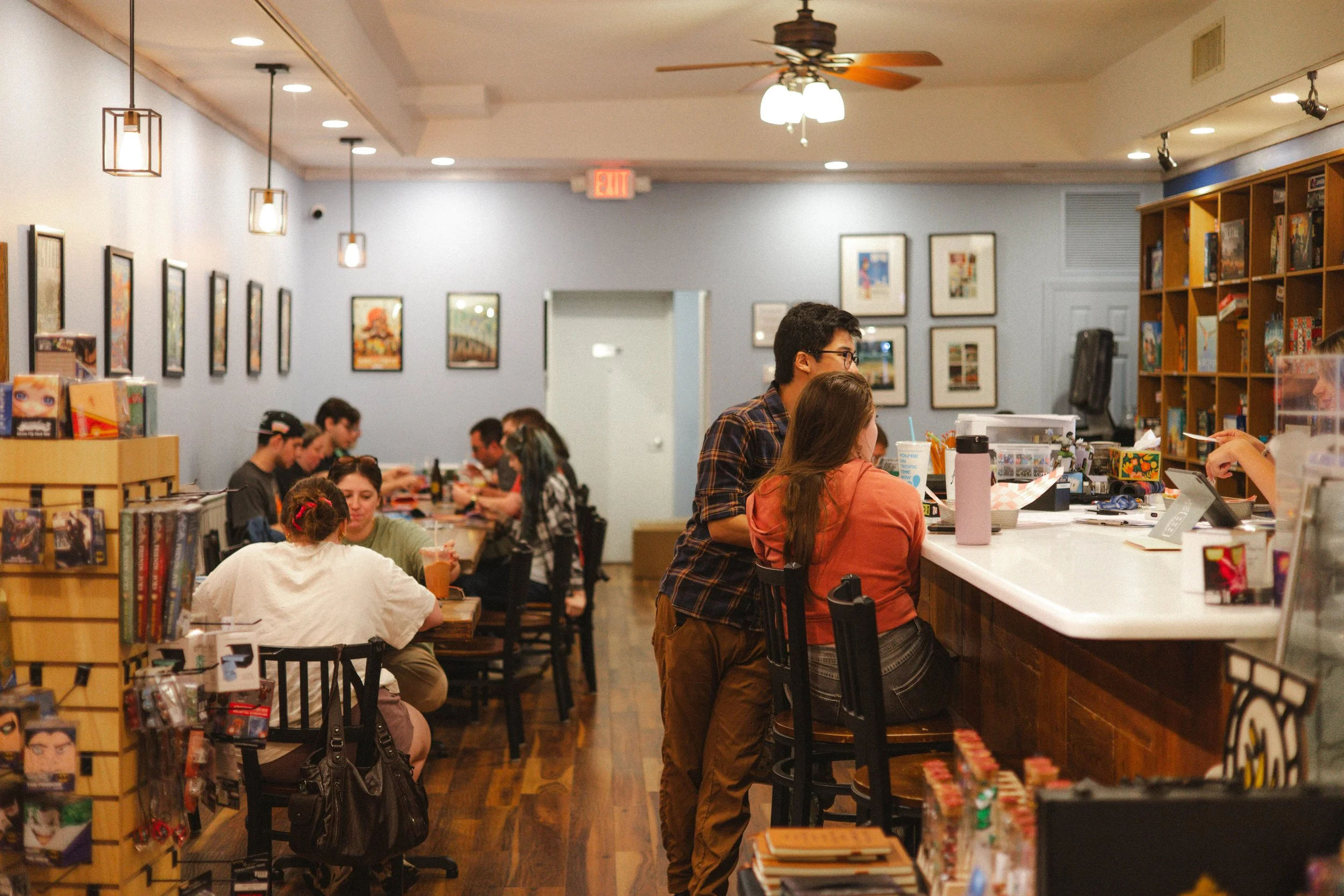 People sitting at a long table inside a bookstore cafe, with shelves of books and artwork on the walls, and standing at the counter, two people engaged in a conversation.