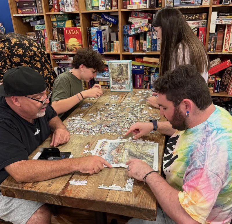 Four people working together on assembling a large, illustrated jigsaw puzzle at a wooden table in a bookstore. Two adults and two children are focused on the puzzle, with shelves of books in the background.