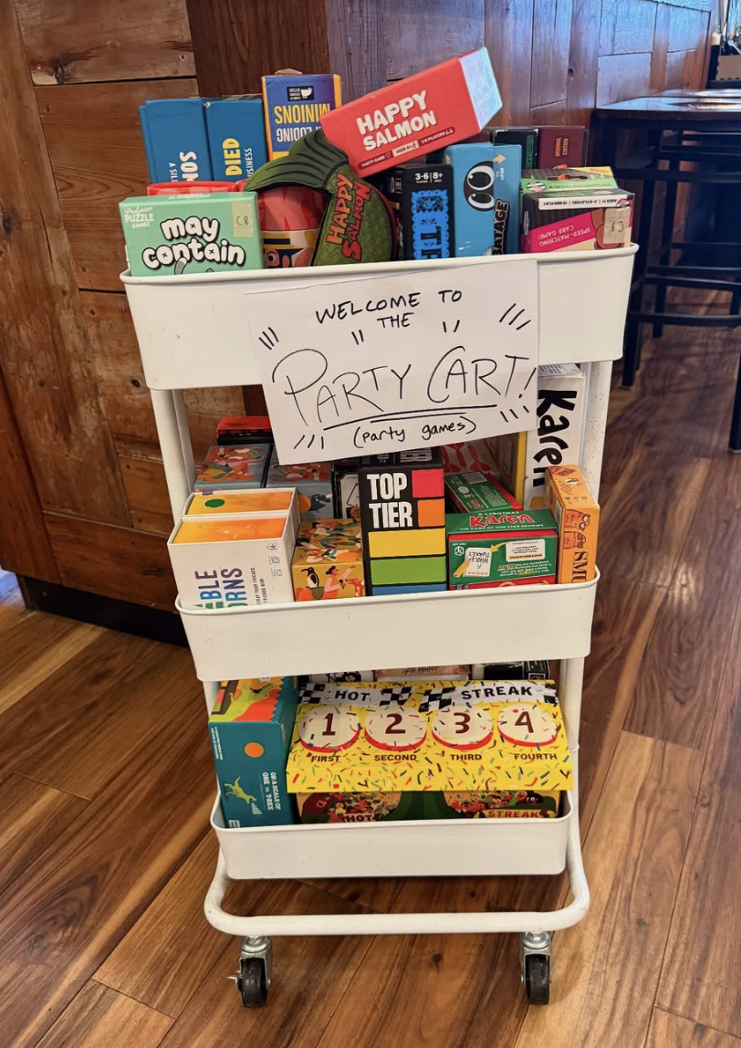 A white three-tiered cart filled with various party games and toys, with a handwritten sign that reads, "Welcome to the Party Art! (party games)." The top shelf has games like Happy Salmon and My Contain, the middle shelf has games like Top Tier and